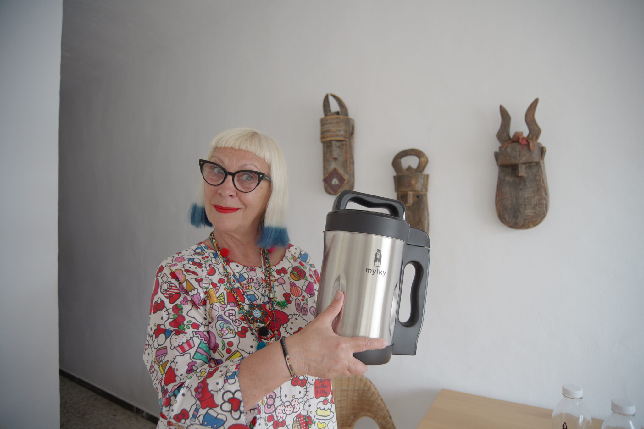 Petra Barkhof holding the Mylky plant milk maker at home, wearing a colorful Hello Kitty print top and signature blond hairstyle with blue tips. Lifestyle portrait for Scimparello Magazine.