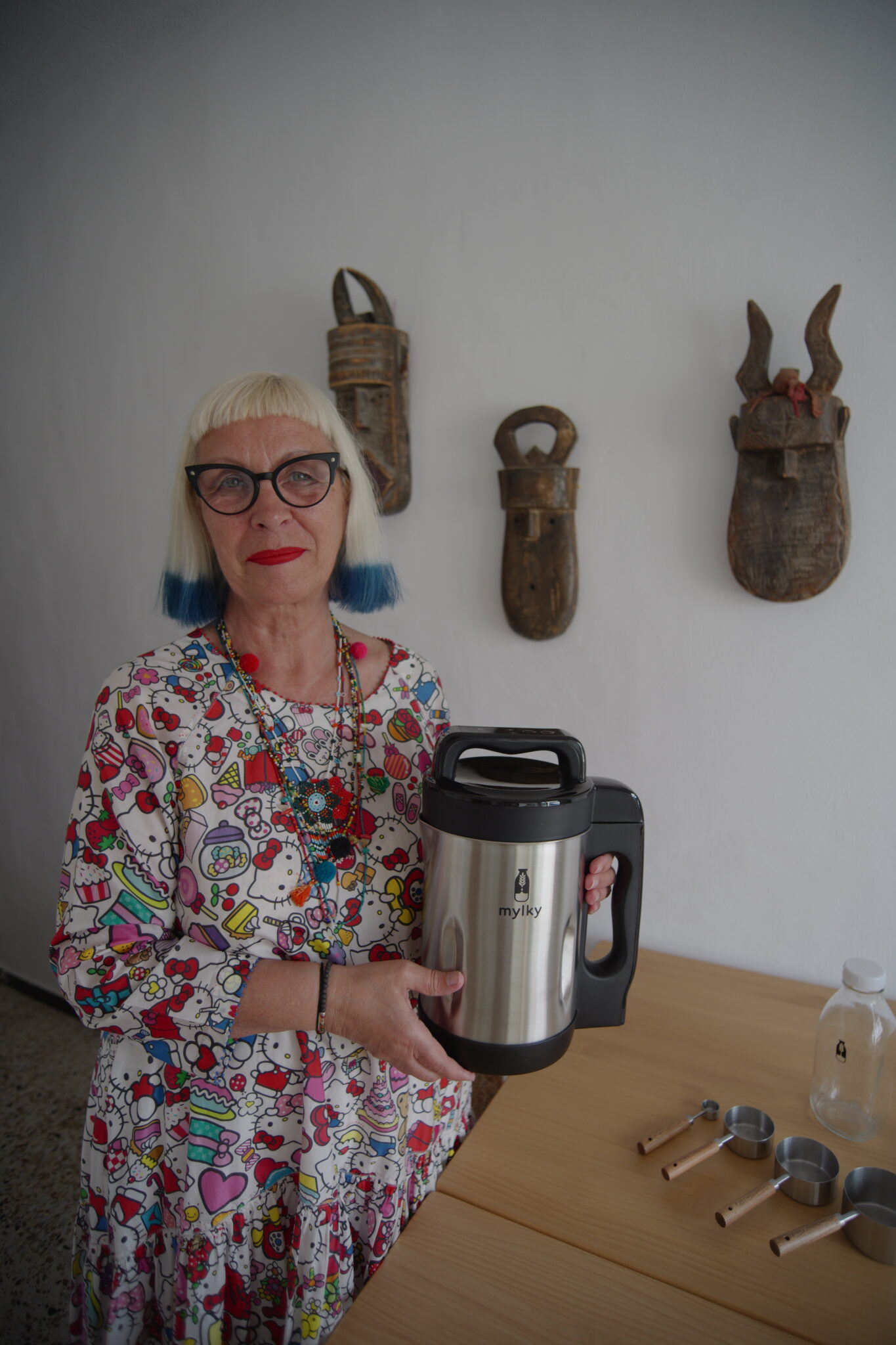Petra Barkhof holding the Mylky plant milk maker at home, wearing a colorful Hello Kitty print top and signature blond hairstyle with blue tips. Lifestyle portrait for Scimparello Magazine.