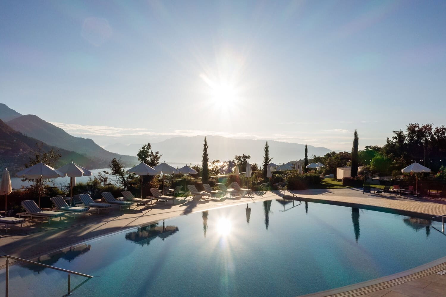 Sunset view over the outdoor pool at A-ROSA Lago di Garda, with white loungers, umbrellas and panoramic mountain views reflected on the water.