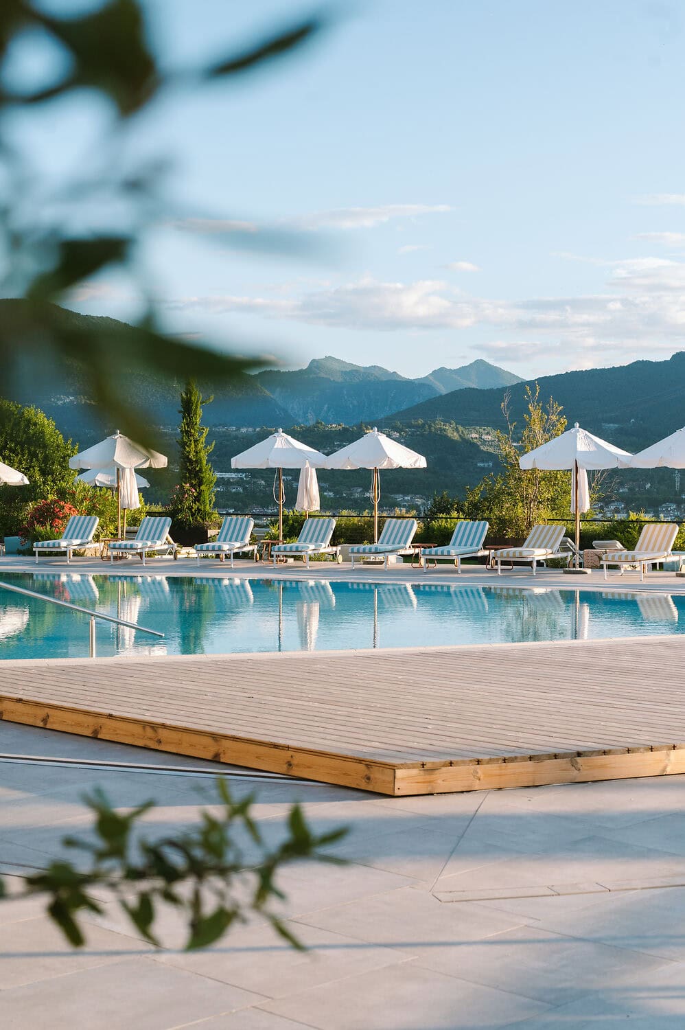 Elegant outdoor swimming pool at A-ROSA Lago di Garda with striped sun loungers, white parasols and scenic views of the surrounding hills.