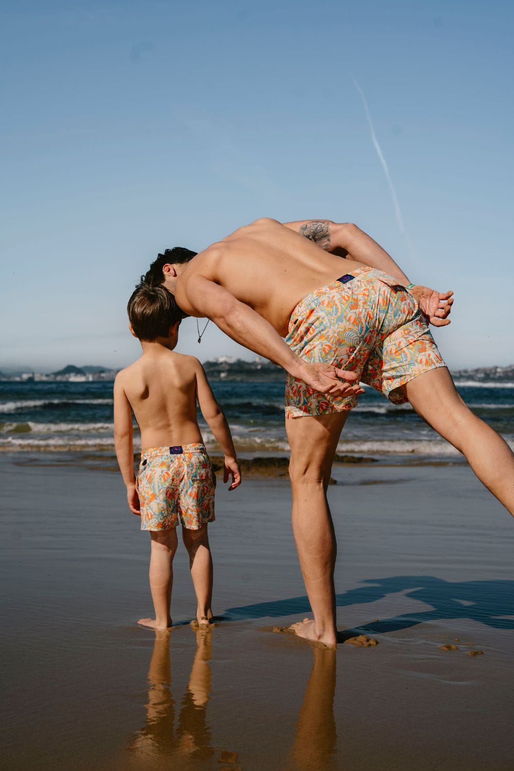 Father and son wearing matching printed swim shorts from Flamenco Maltés SS26 walk along the shoreline together, capturing the relaxed spirit of mini me beachwear and contemporary Mediterranean family style.