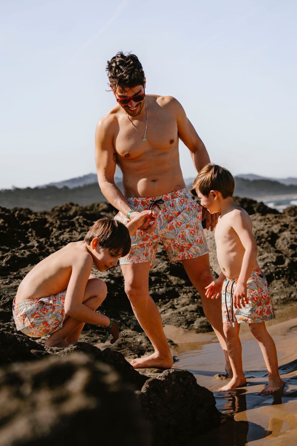 Father and son wearing matching printed swim shorts from Flamenco Maltés SS26 walk along the shoreline together, capturing the relaxed spirit of mini me beachwear and contemporary Mediterranean family style.