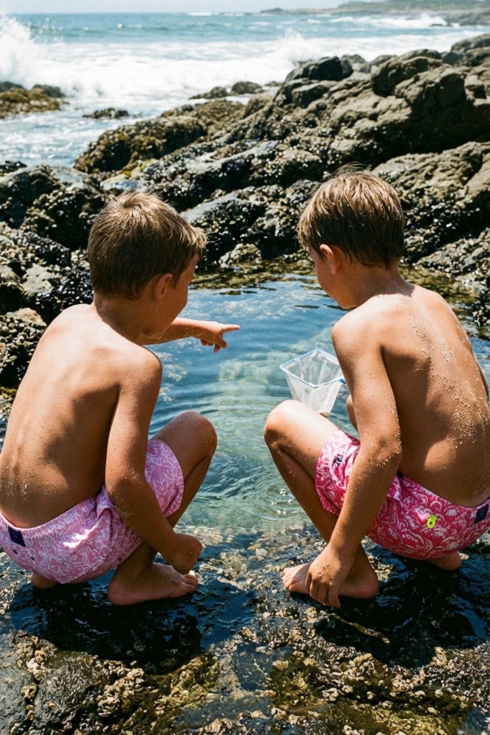 Two boys wearing pink patterned swim shorts discover sea life in a natural rock pool, embodying the adventurous and playful mood of Flamenco Maltés kids swimwear for SS26.