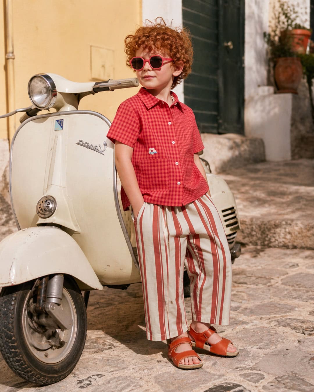 Curly-haired child wearing red gingham shirt, striped beige and red trousers, and red sandals, leaning against a vintage cream scooter, with pink sunglasses, in a sunny Mediterranean street setting.