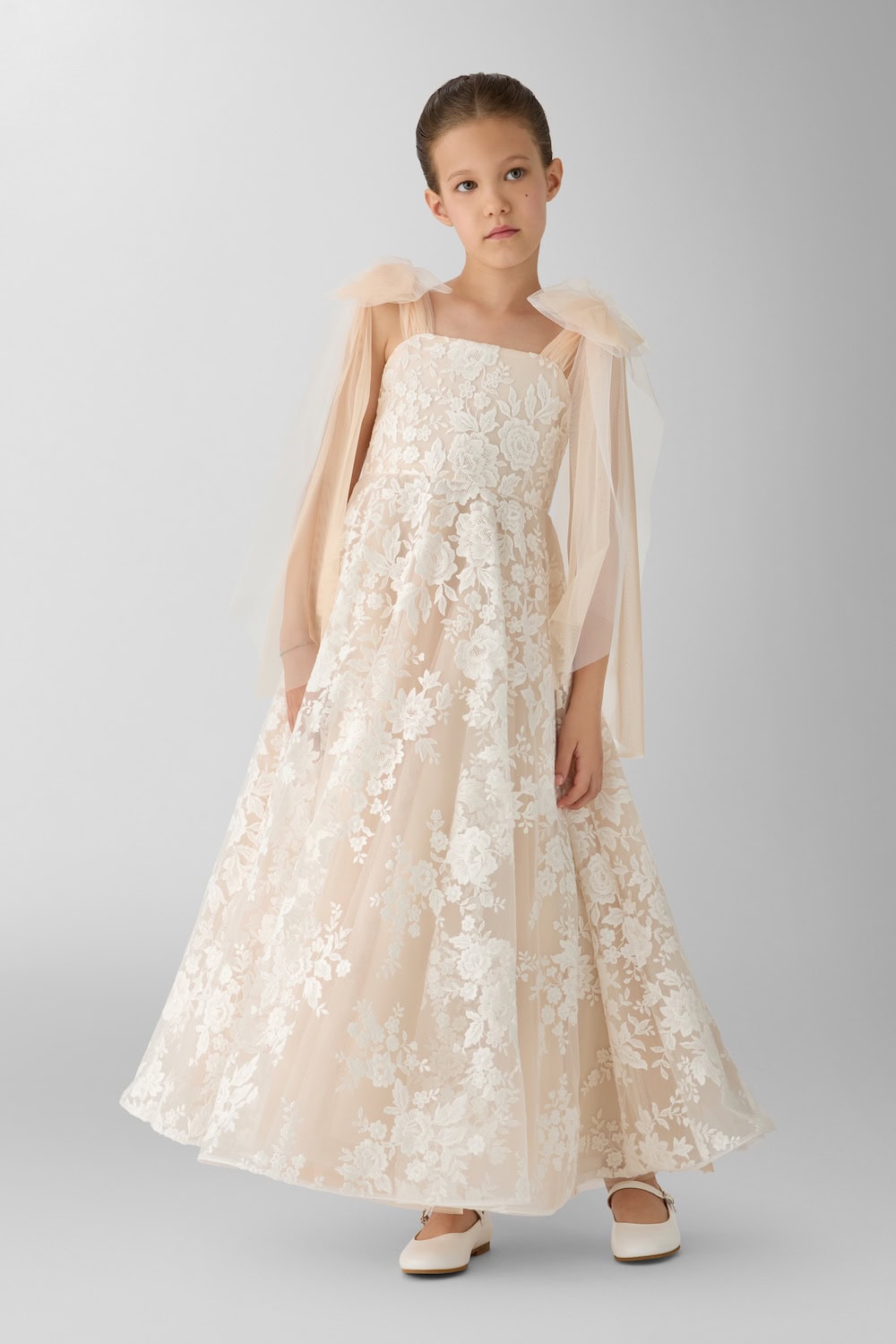 Girl in a blush-toned lace gown with oversized tulle shoulder bows and full floral skirt in a minimalist studio setting.