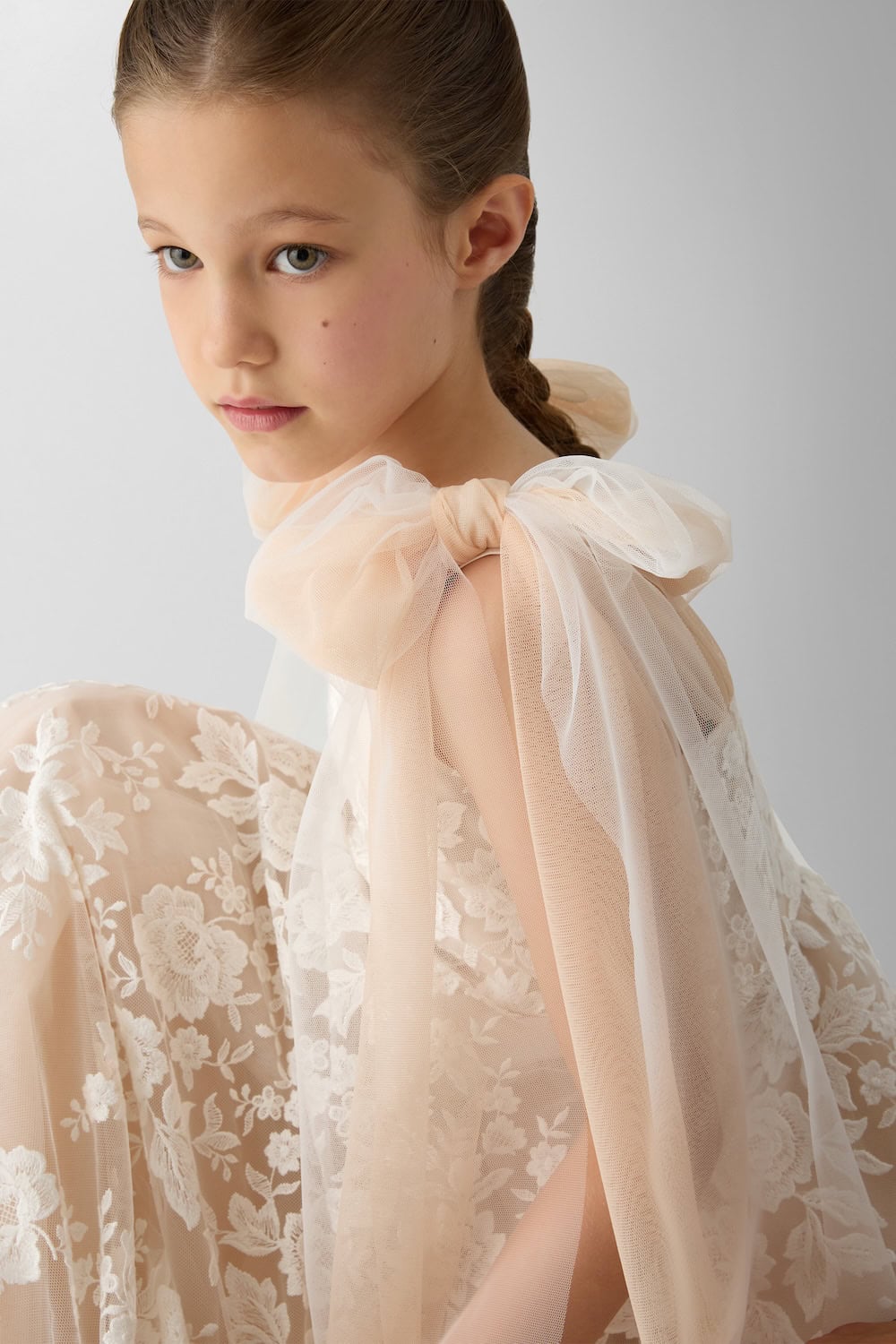 Girl in a blush-toned lace gown with oversized tulle shoulder bows and full floral skirt in a minimalist studio setting.