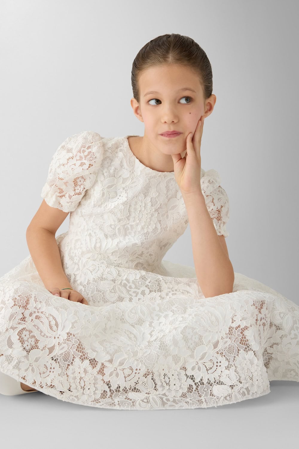 Girl sitting in a full white lace dress with puff sleeves and intricate floral texture photographed against a neutral background.
