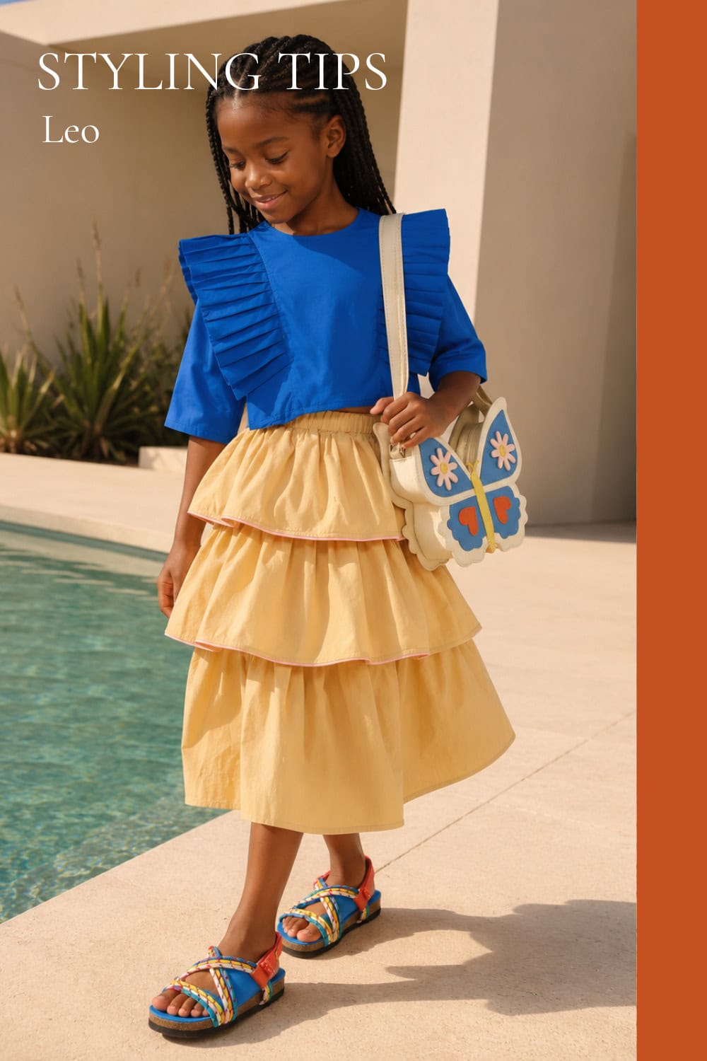 Young girl wearing a bright blue pleated ruffle top with a layered pale yellow skirt, colorful sandals and a butterfly-shaped bag while walking beside a pool in a modern sunny villa setting.