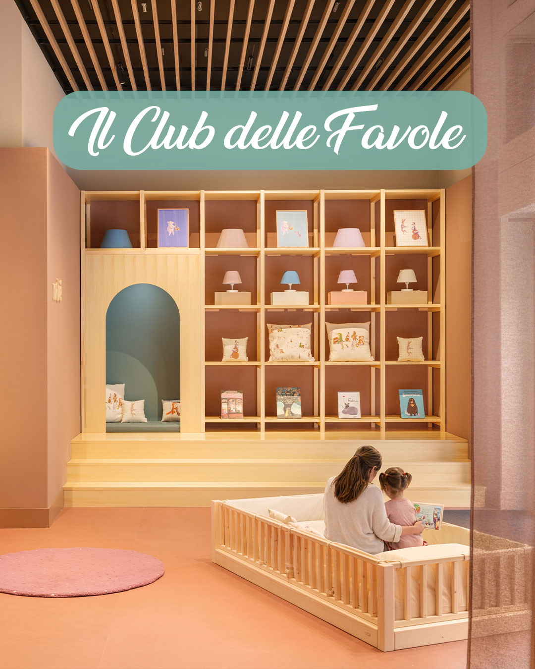 Warm pastel-toned interior of Nabè’s Milan flagship store featuring Montessori-inspired children’s furniture, soft pink flooring, wooden shelving and a mother reading a book with her child on a low bed during “Il Club delle Favole”.
