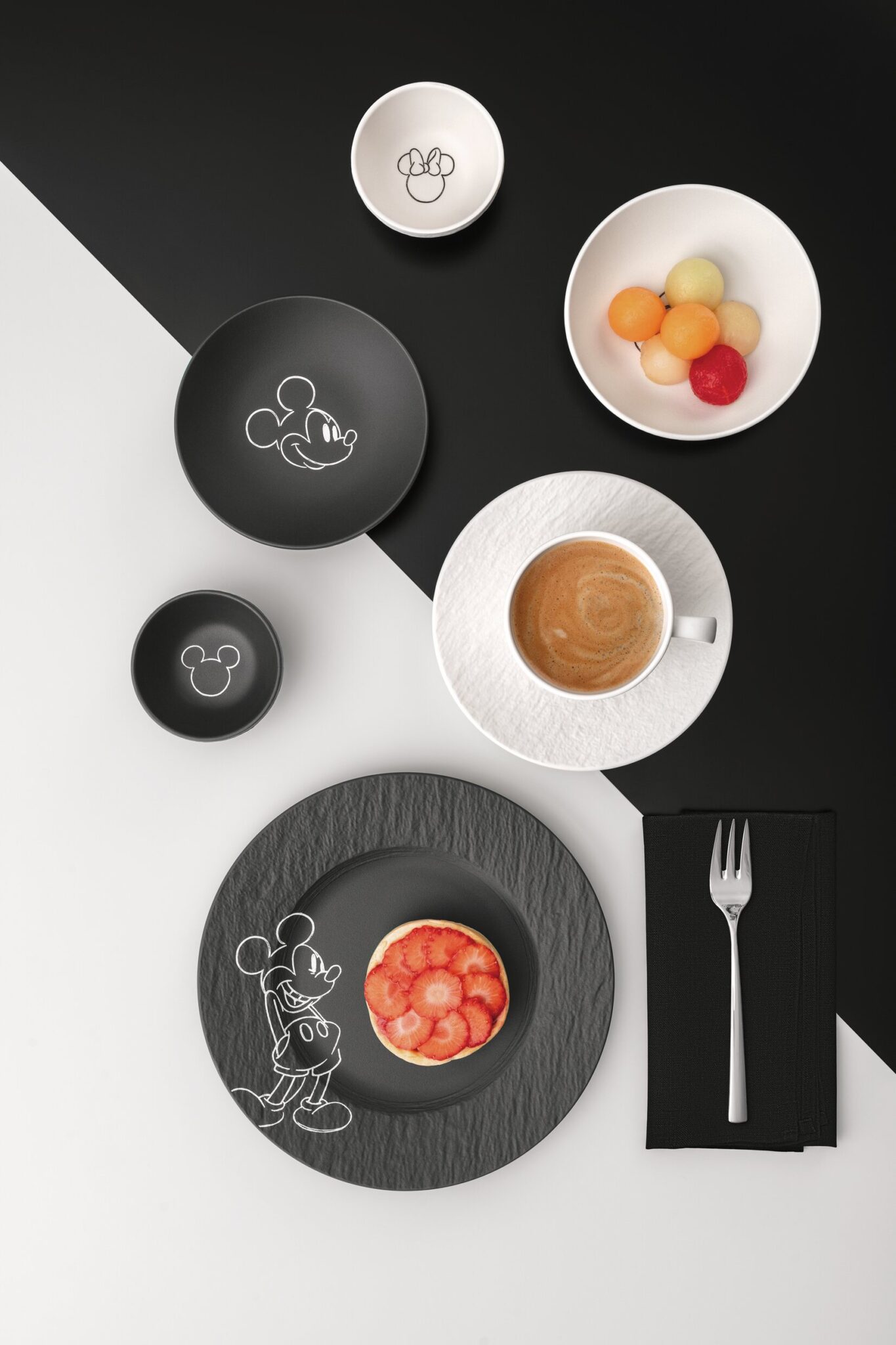 Minimalist breakfast table setting from the Manufacture – Mickey Mouse collection with textured black and white porcelain, espresso cup, dessert plate, fruit bowl, and illustrated Mickey Mouse details in a modern black-and-white composition.