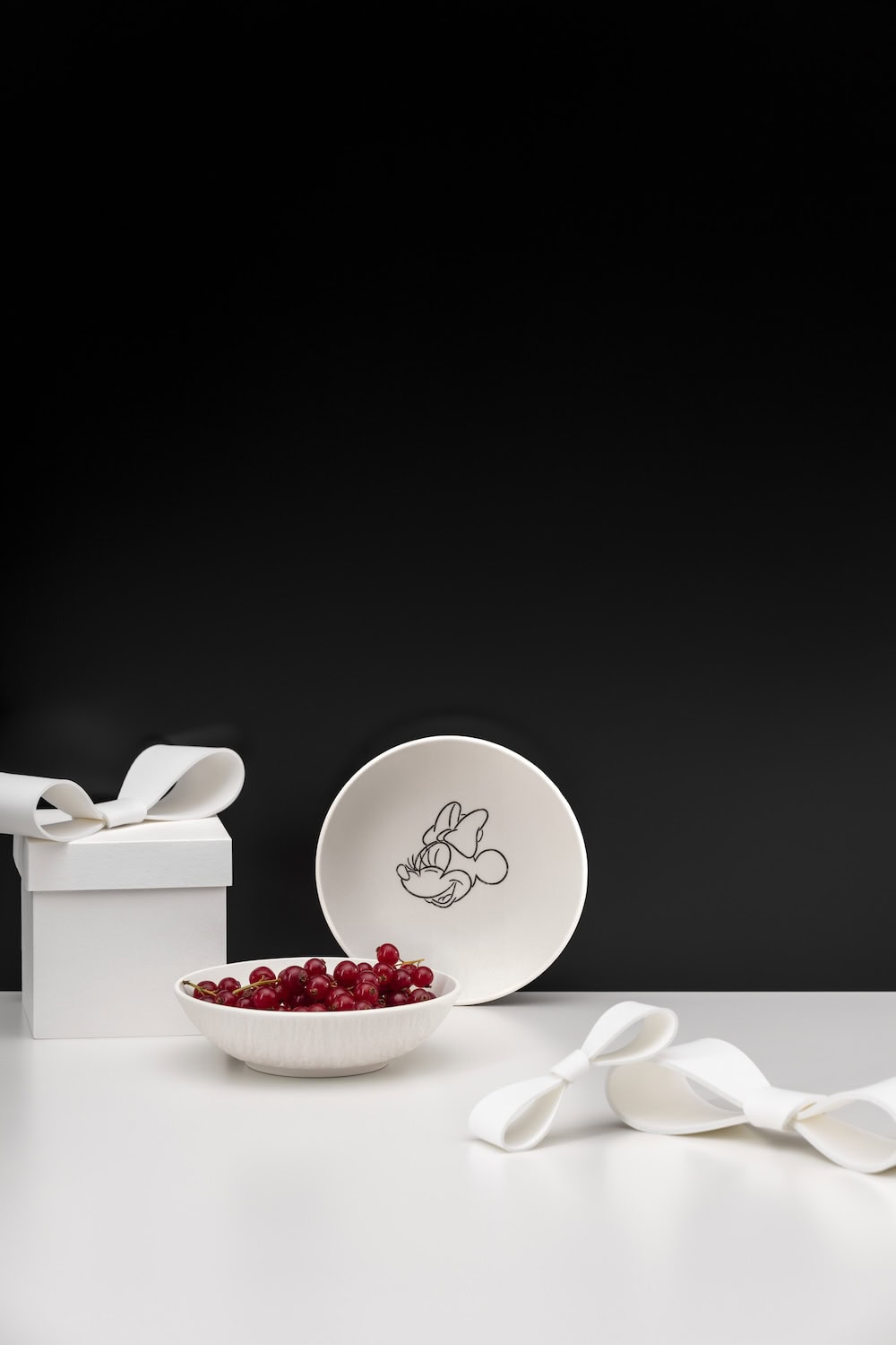 White porcelain bowl from the Manufacture – Mickey Mouse collection decorated with a sketch-style illustration of Minnie Mouse, styled with red currants and monochrome gift boxes against a black-and-white background.