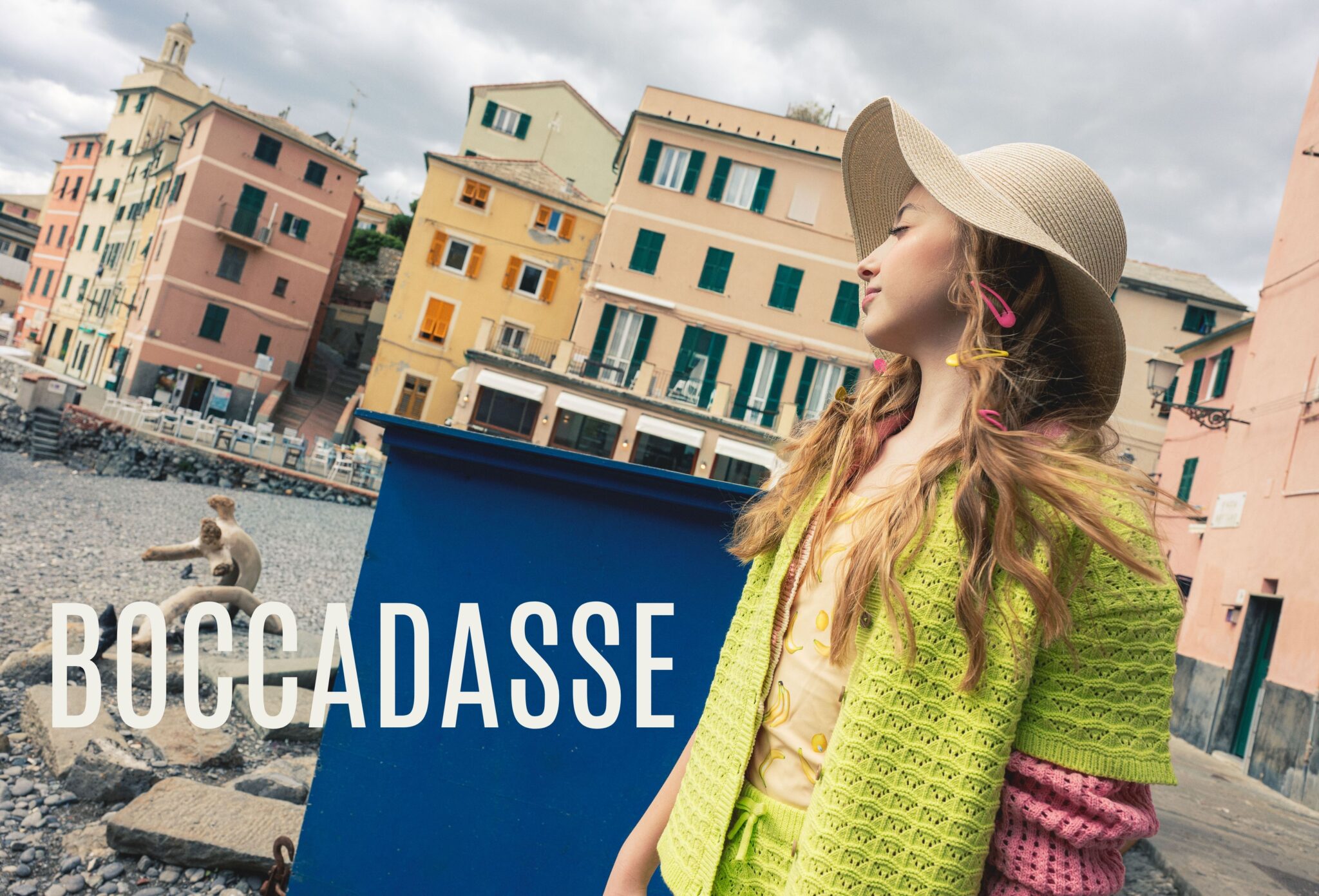 Girl wearing a straw hat and colorful summer knitwear in Boccadasse, Liguria, with pastel seaside houses in the background, photographed by Ilaria & Silvia Cianciaruso for Scimparello Magazine.