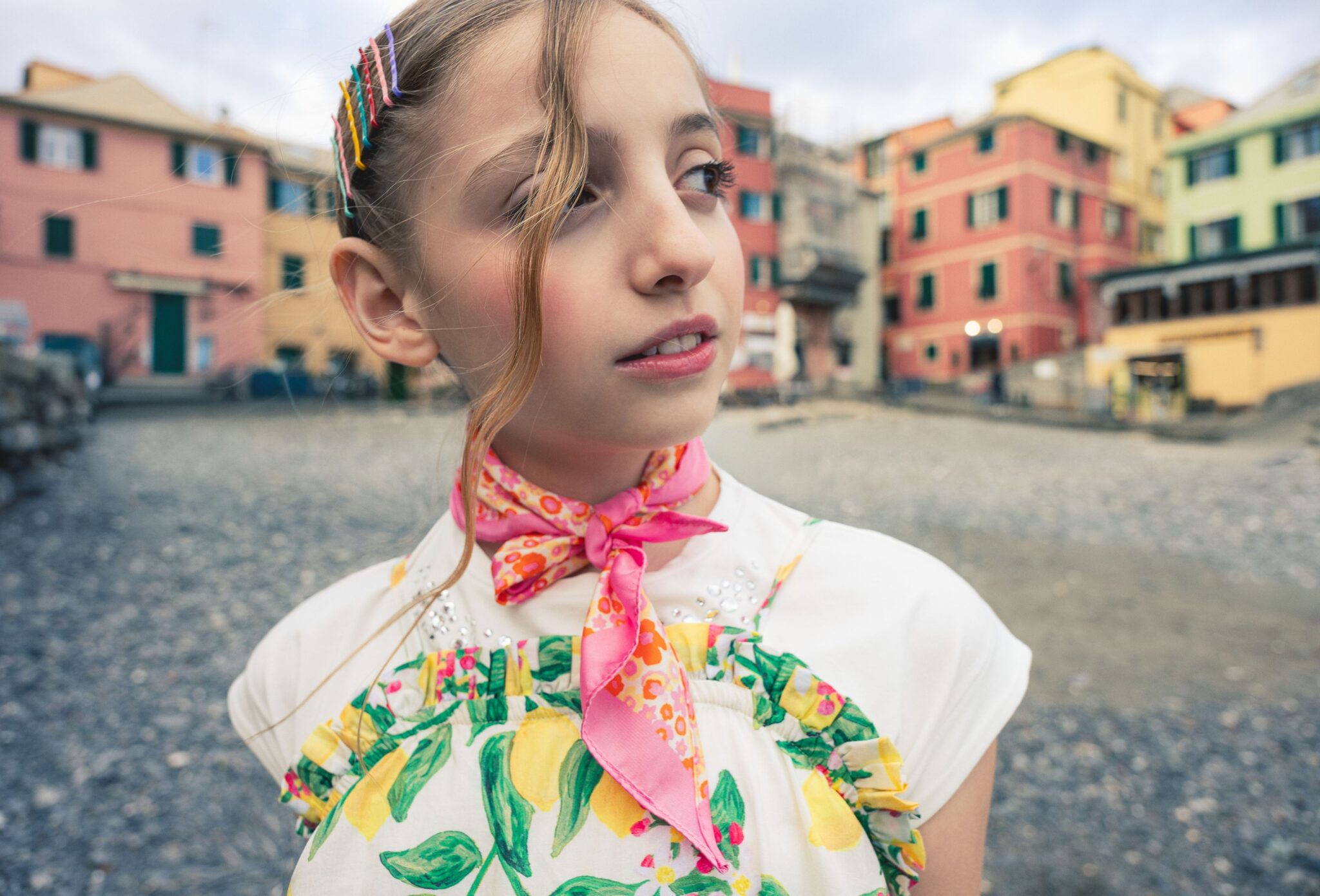 Close-up portrait of a girl wearing a lemon-print summer dress, pink neck scarf and colorful hair clips on the beach in Boccadasse, Liguria, photographed by Ilaria & Silvia Cianciaruso for Scimparello Magazine.
