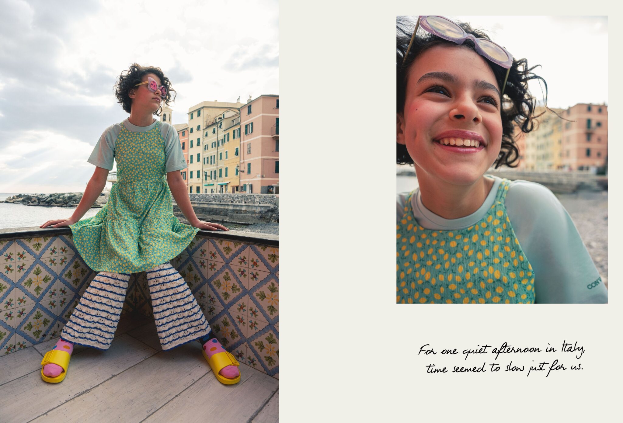 Girl wearing a turquoise polka dot dress over striped trousers, pink socks, yellow sandals and sunglasses in Boccadasse, Liguria, with pastel seaside houses behind her, photographed by Ilaria & Silvia Cianciaruso for Scimparello Magazine.