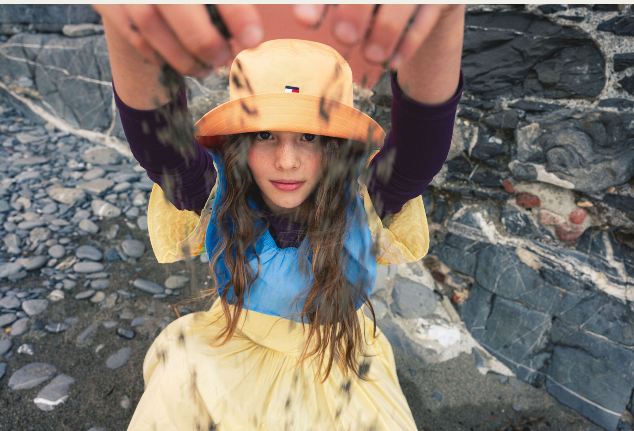 Girl in a yellow summer dress and orange bucket hat reaching toward the camera on the rocky beach in Boccadasse, Liguria, photographed by Ilaria & Silvia Cianciaruso for Scimparello Magazine.