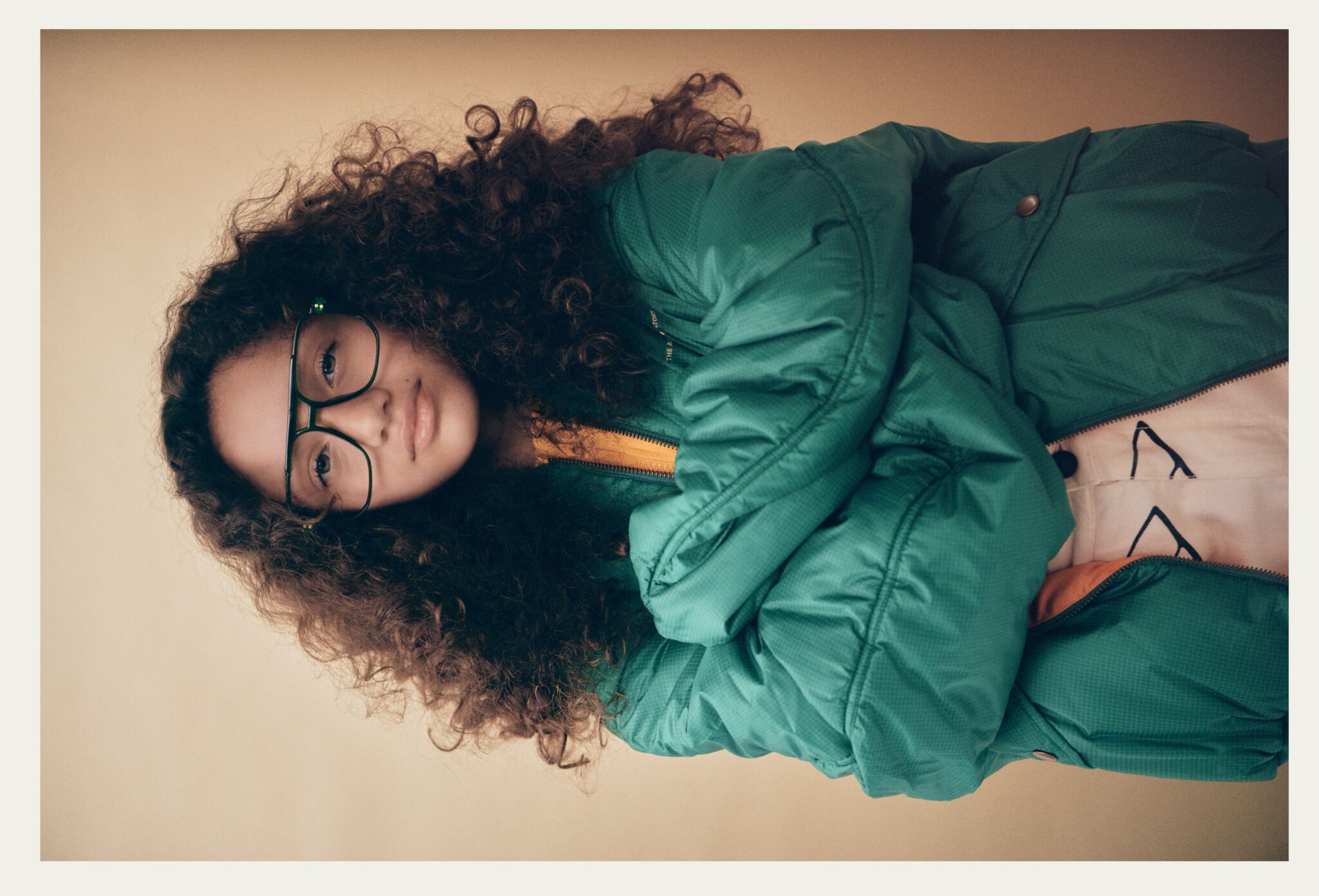 Portrait of a child with curly hair and green glasses wearing an oversized green padded jacket in Faces, a kids fashion editorial photographed by Franck Malthierry for Scimparello Magazine.