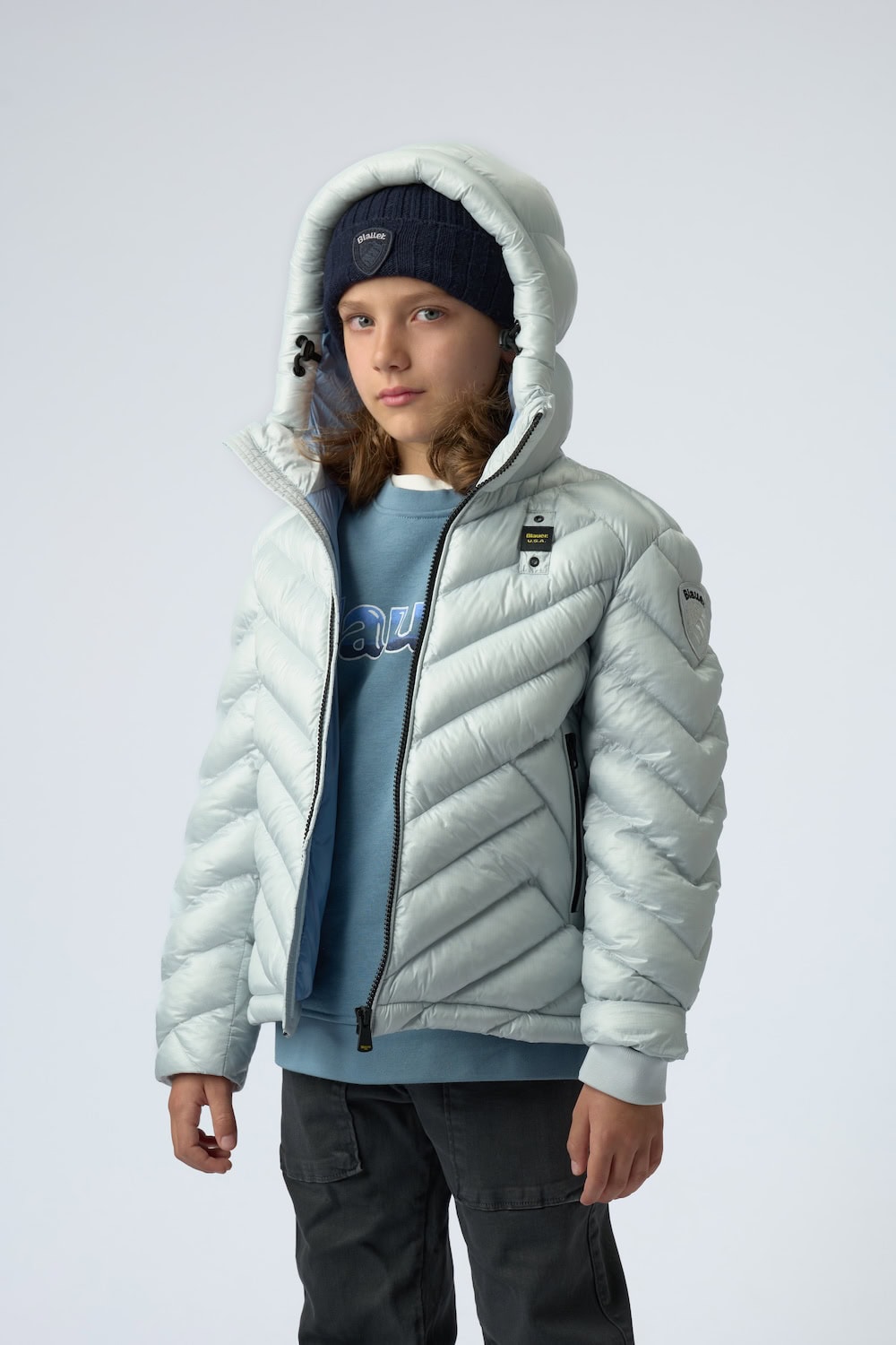 Boy wearing a light ice-blue quilted down jacket with hood, pale blue sweatshirt and navy knit beanie from the Blauer Junior FW26/27 collection.