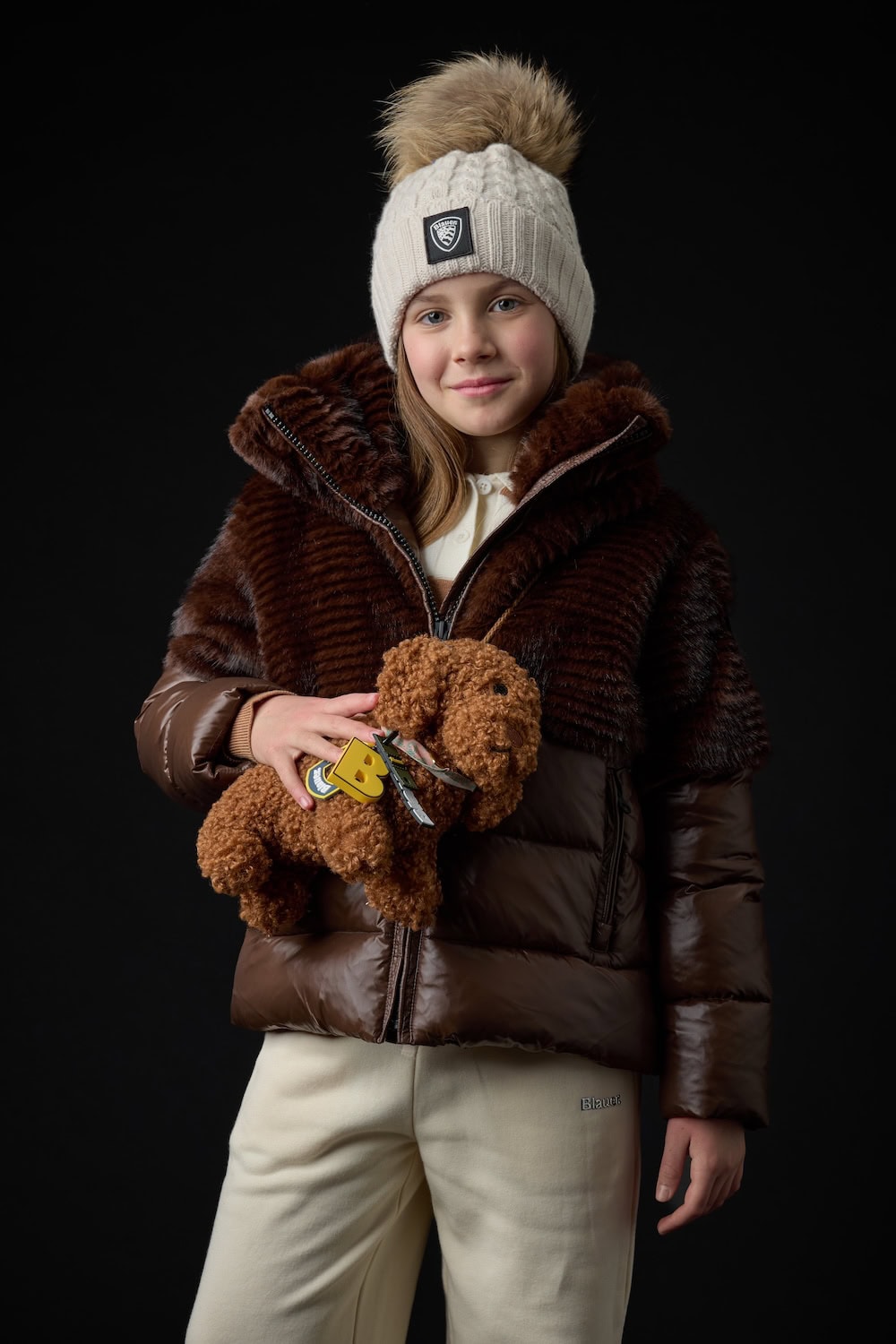 Girl wearing a brown mixed-texture winter jacket with faux-fur details, cream knit hat and matching trousers, holding a teddy dog from the Blauer Junior FW26/27 collection.