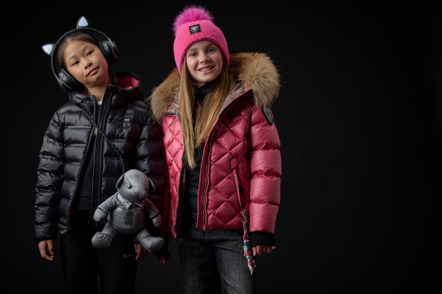 Two girls wearing glossy puffer jackets in black and burgundy with faux-fur trims, styled with playful accessories from the Blauer Junior FW26/27 collection.