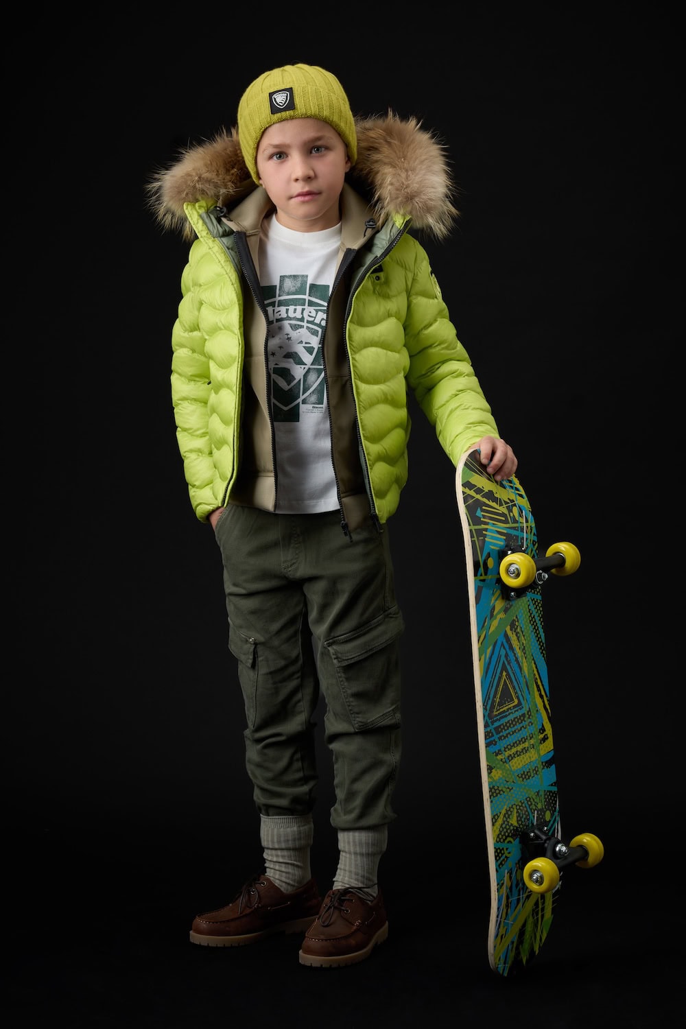 Boy wearing a lime green puffer jacket with fur-trimmed hood, cargo trousers and knit beanie, holding a skateboard from the Blauer Junior FW26/27 collection.