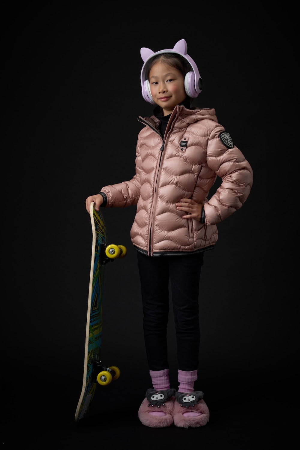 Girl wearing a blush pink quilted puffer jacket, black leggings and oversized headphones, posing with a skateboard from the Blauer Junior FW26/27 collection.