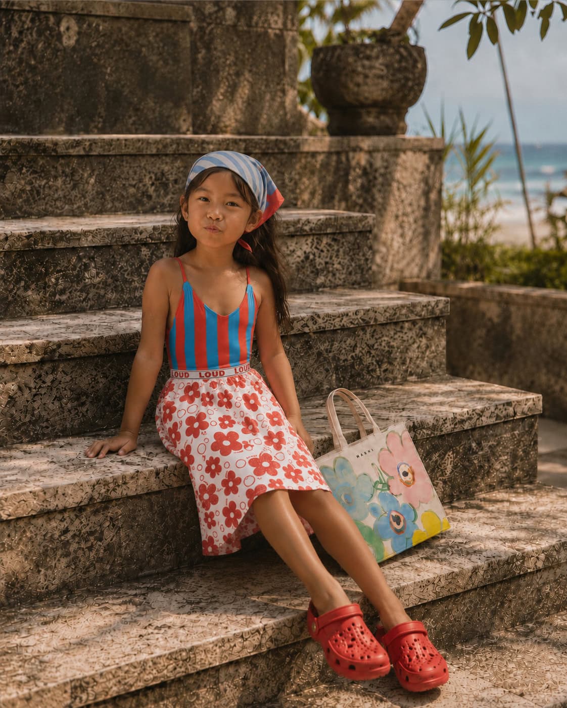 Girl wearing a striped bathing suit with a red floral skirt, striped headscarf, red clogs and floral bag on tiled seaside steps in Bali for a Styling Tip on floral skirts, Scimparello Magazine.