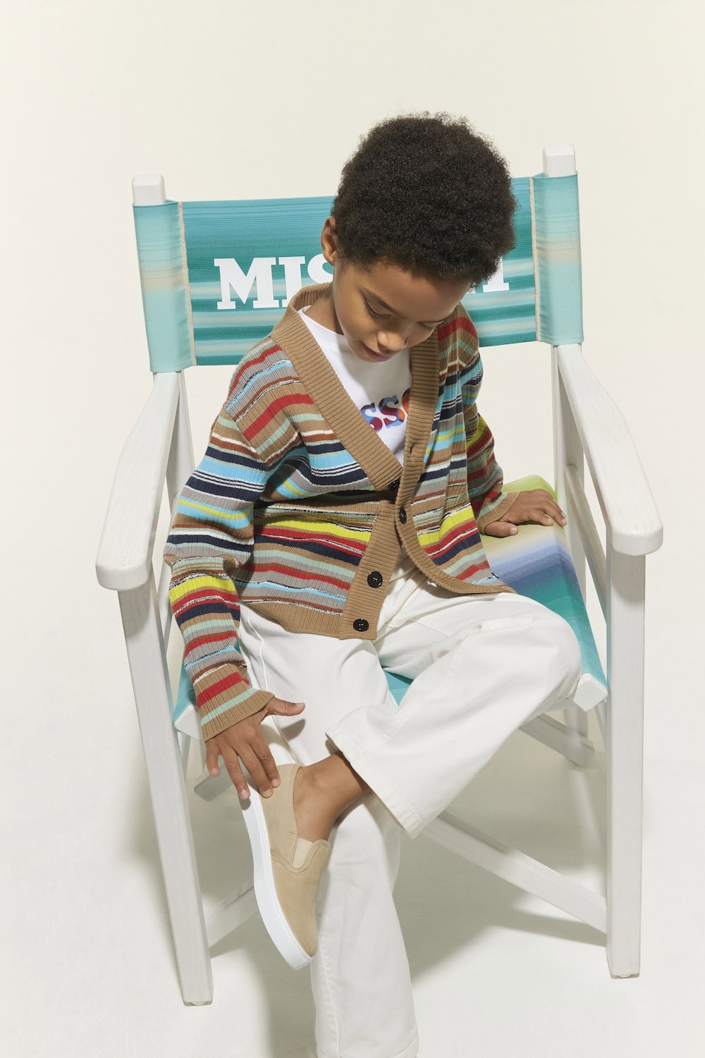 Boy seated in a beach chair wearing a striped Missoni Kids SS26 knitted cardigan, white trousers and casual summer shoes, showcasing the collection's relaxed resort aesthetic.