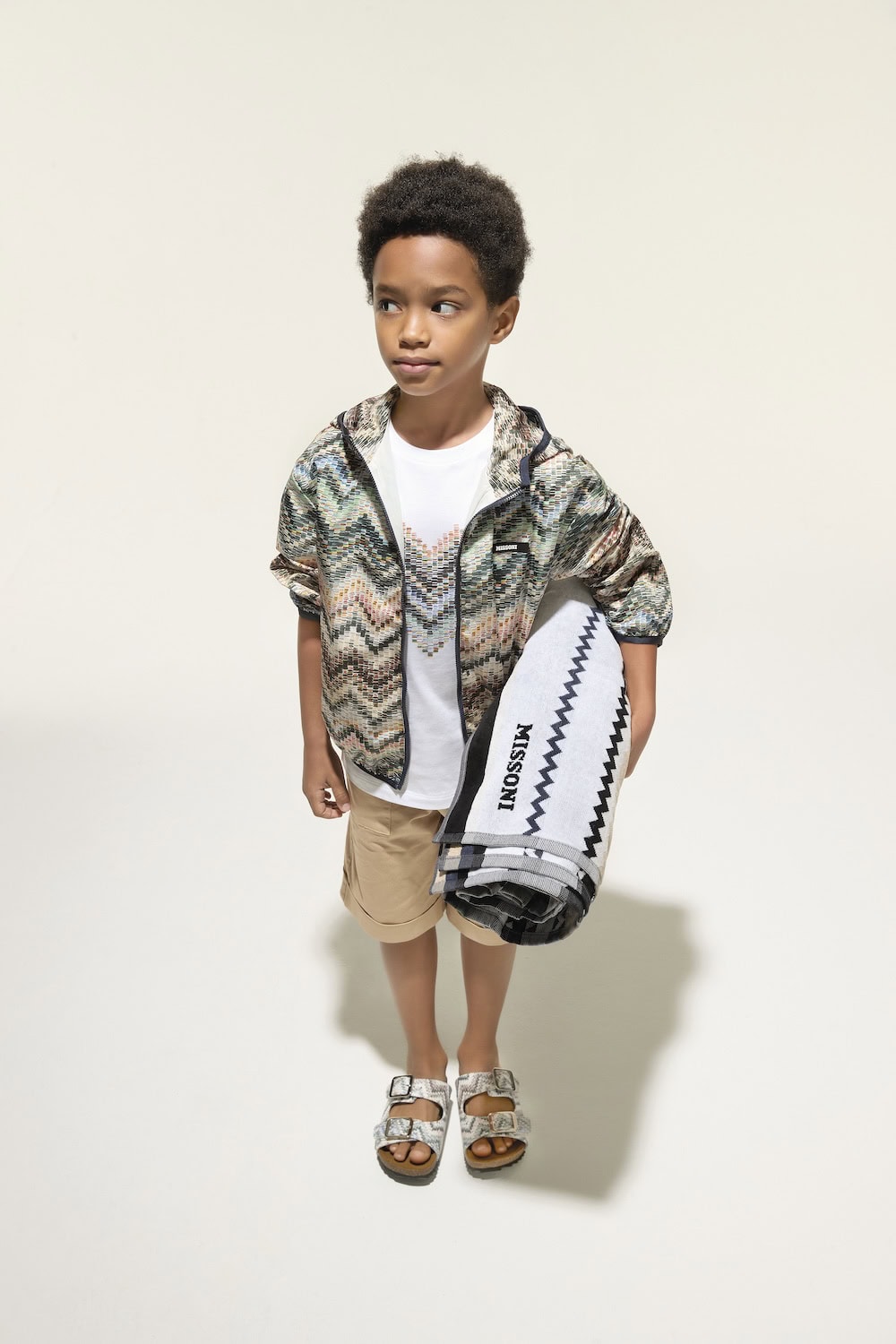 Boy wearing a Missoni Kids SS26 zigzag-print lightweight jacket, beige shorts and logo T-shirt, holding a Missoni beach towel for a contemporary seaside-inspired look.