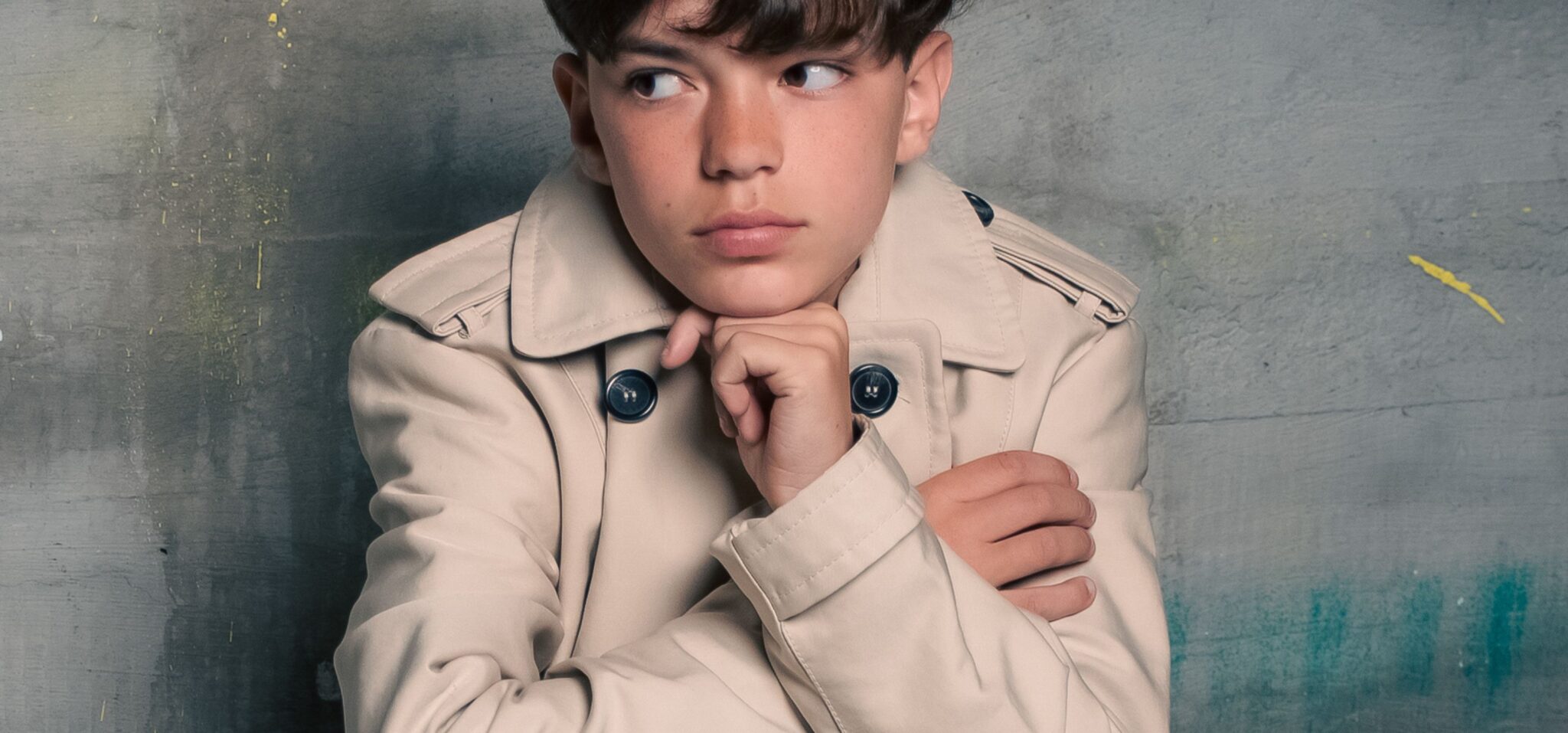 Boy sitting backwards on a wooden chair in a beige trench coat, posing against a textured grey wall in the Friends Forever kids fashion editorial photographed by Danique Daemen, Scimparello Magazine.
