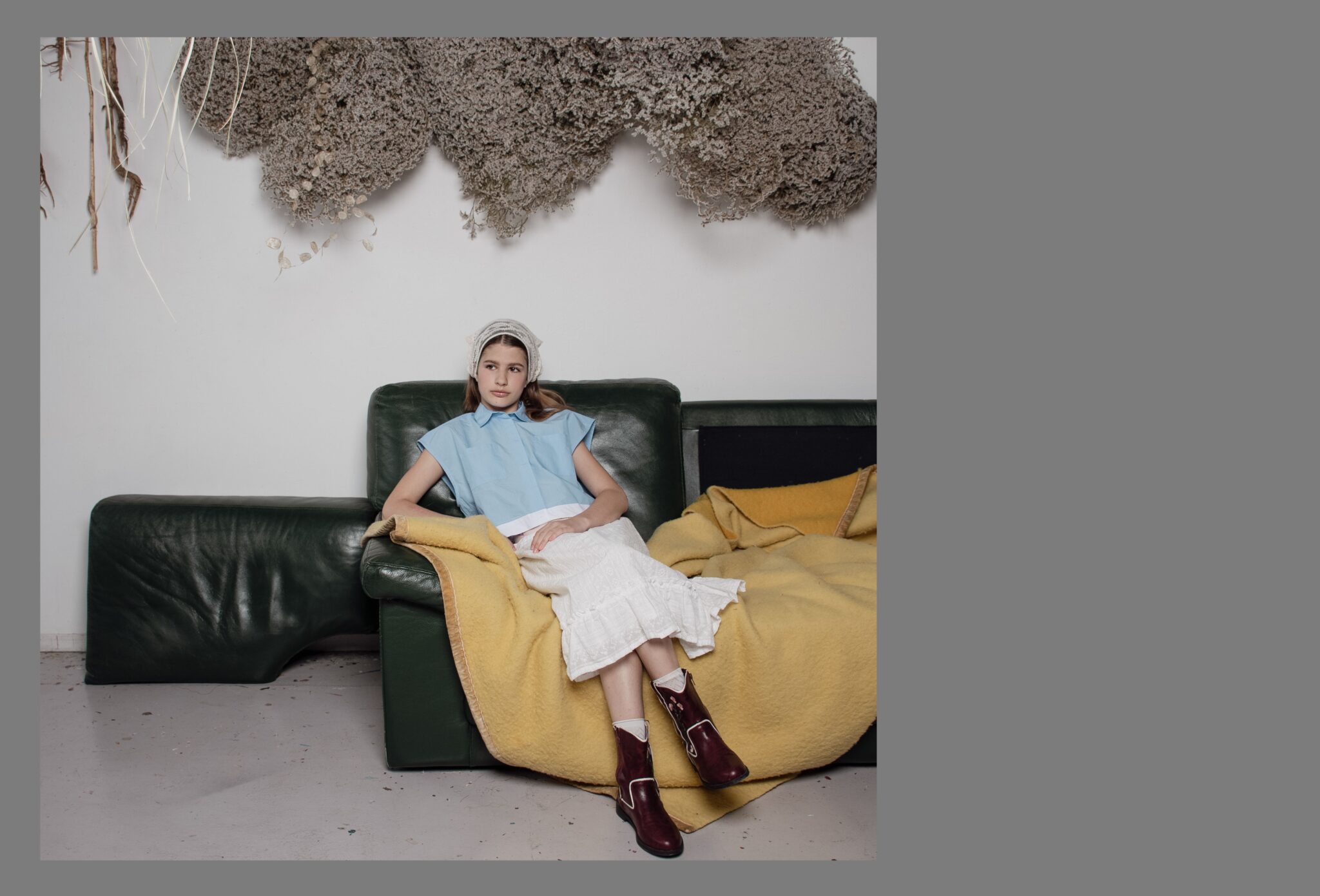 Child sitting on a dark green sofa with a yellow blanket, wearing a pale blue top, white skirt, headscarf, and burgundy boots in the Friends Forever kids fashion editorial photographed by Danique Daemen, Scimparello Magazine.