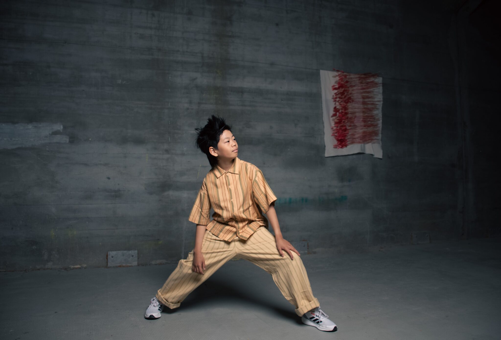 Boy posing in a wide stance against a grey industrial wall, wearing a striped beige shirt and matching trousers in the Friends Forever kids fashion editorial photographed by Danique Daemen, Scimparello Magazine.