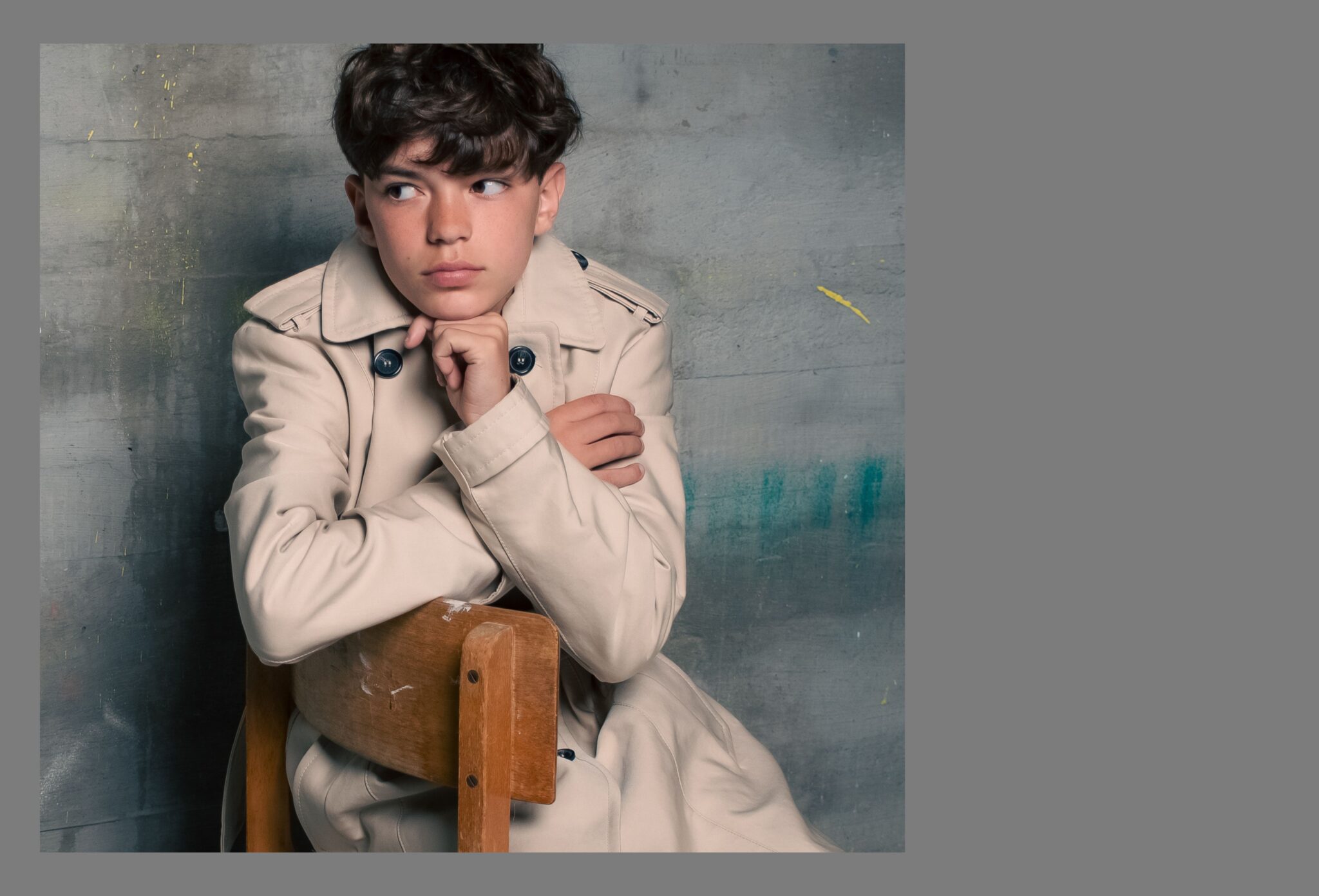 Boy sitting backwards on a wooden chair in a beige trench coat, posing against a textured grey wall in the Friends Forever kids fashion editorial photographed by Danique Daemen, Scimparello Magazine.