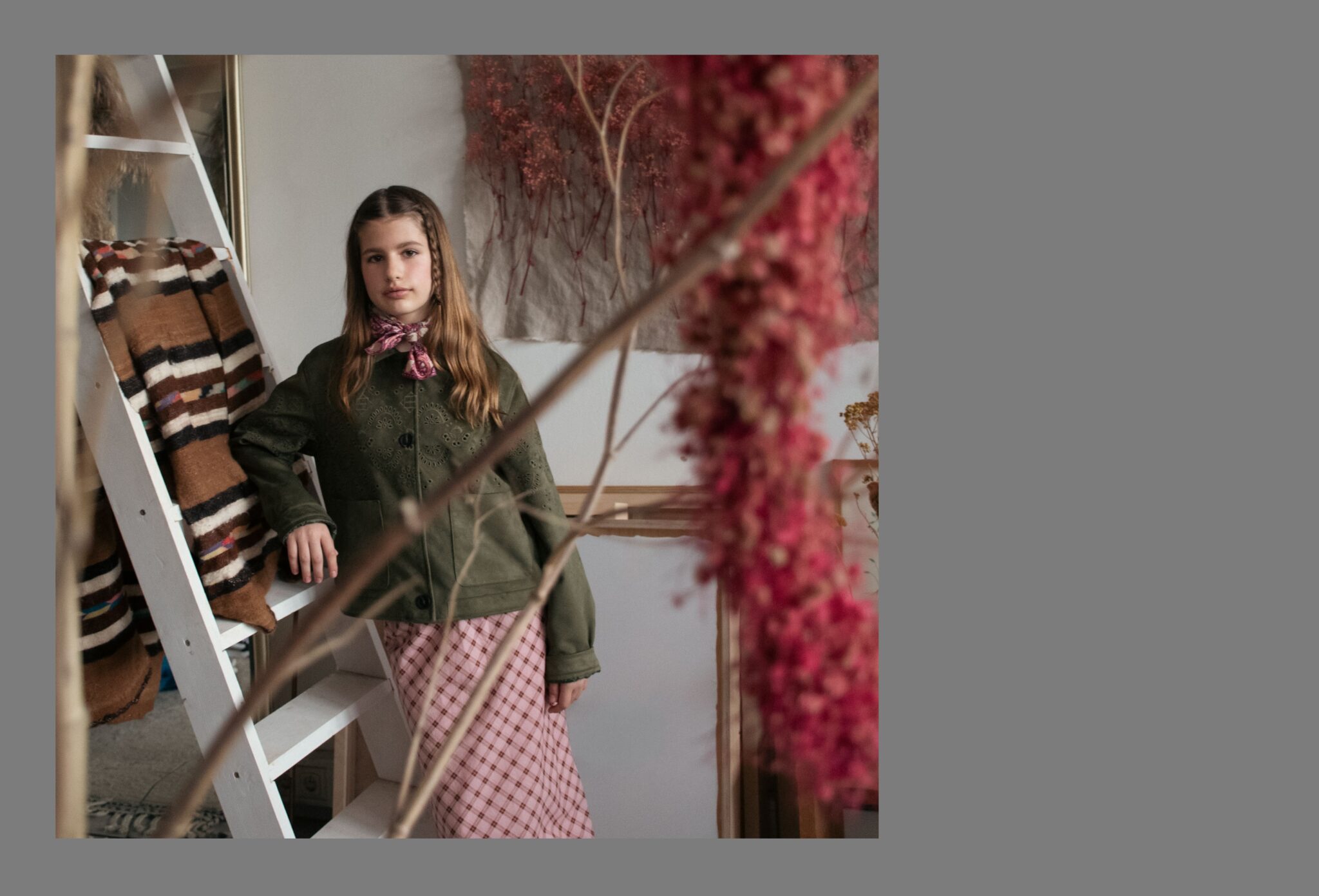 Girl standing in a floral studio wearing an olive green jacket, pink check skirt, and printed neck scarf, framed by dried flowers in the Friends Forever kids fashion editorial photographed by Danique Daemen, Scimparello Magazine.