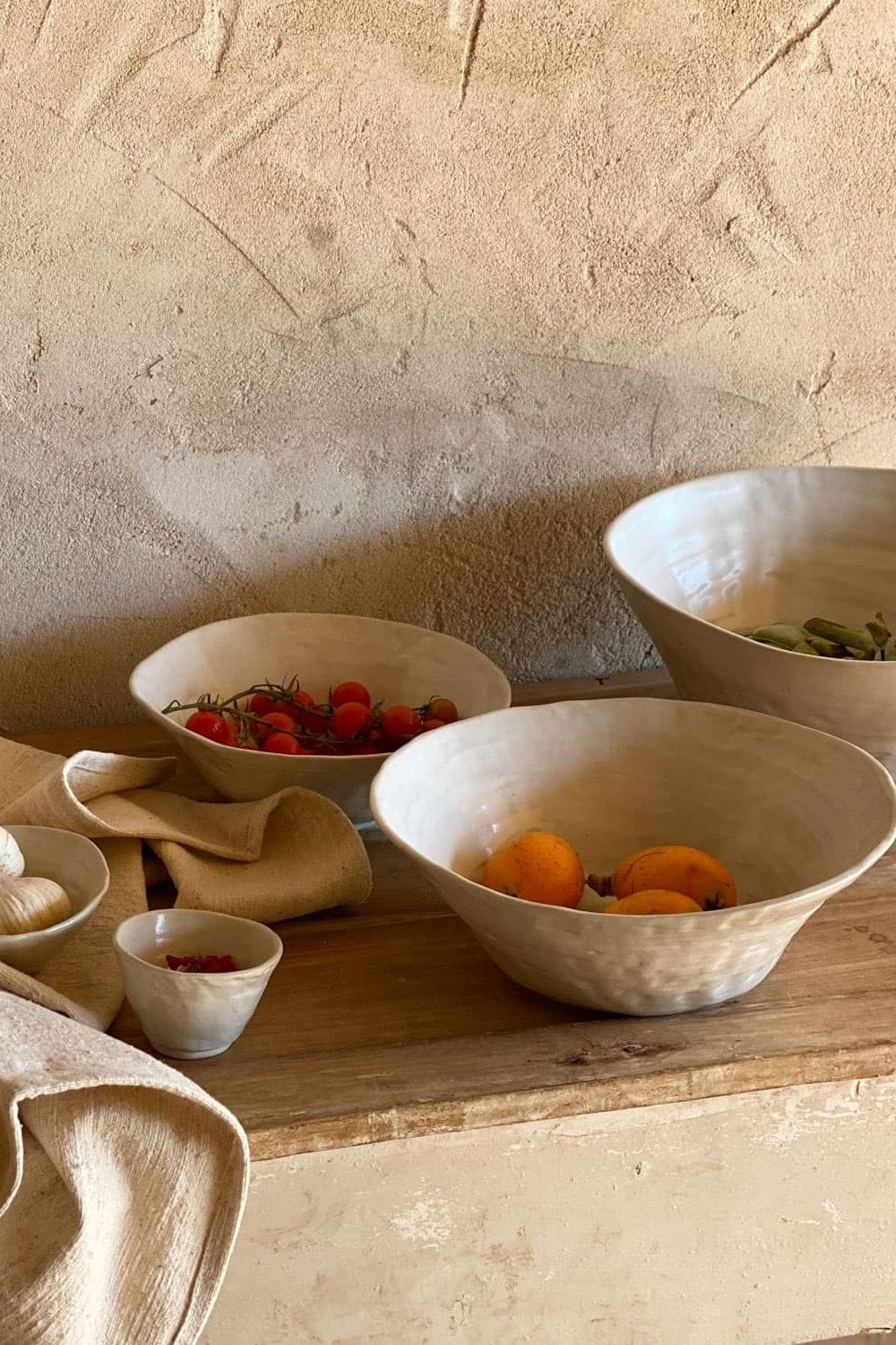 Handcrafted ceramic bowls by N°74 displayed on a rustic wooden table with garlic, tomatoes, apricots and natural linen textiles in a warm Mediterranean-inspired setting.