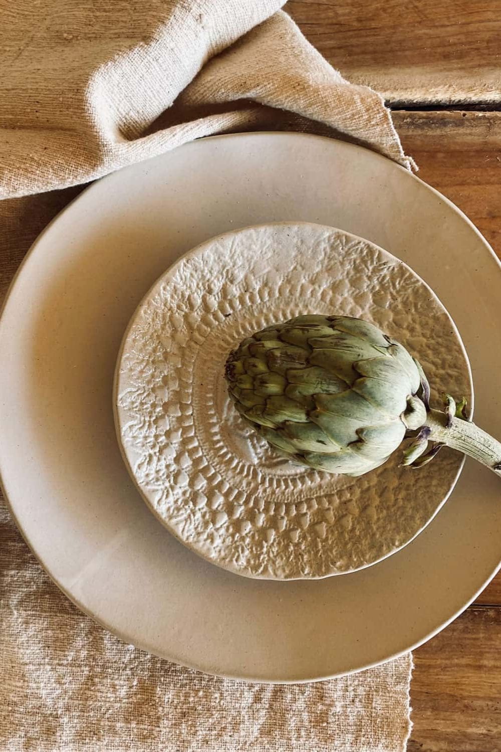 Textured handcrafted ceramic plate and bowl by N°74 featuring an artichoke centerpiece on natural linen fabric and rustic wooden table.