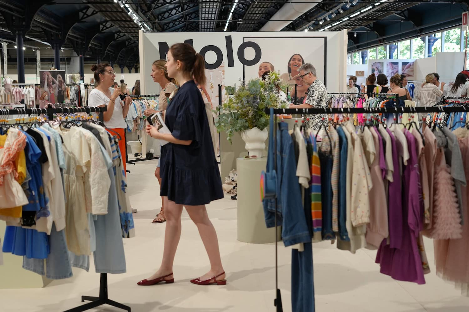 Playtime Paris exhibition floor featuring children's fashion collections displayed on clothing racks, with buyers and industry professionals exploring the latest kidswear trends in a bright trade show setting.