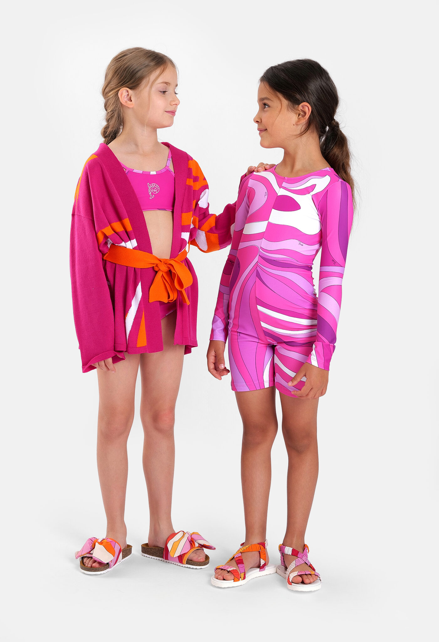 Two girls wearing Pucci Junior SS26 resort looks featuring vibrant pink, orange and lilac Marmo prints, including a knitted cardigan, swimwear and a long-sleeved printed swimsuit.