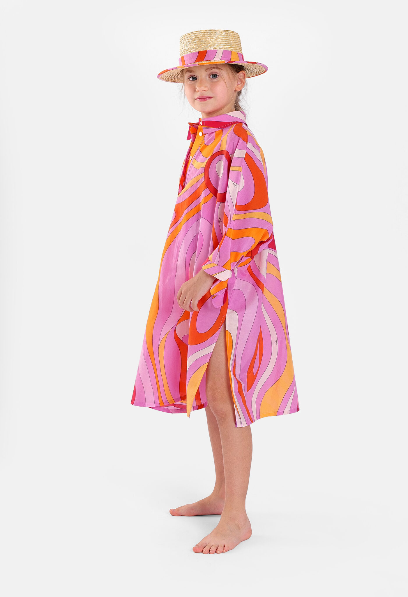 Girl wearing a flowing Pucci Junior SS26 shirt dress featuring the signature Marmo print in shades of pink, orange and lilac, styled with a coordinating straw hat.