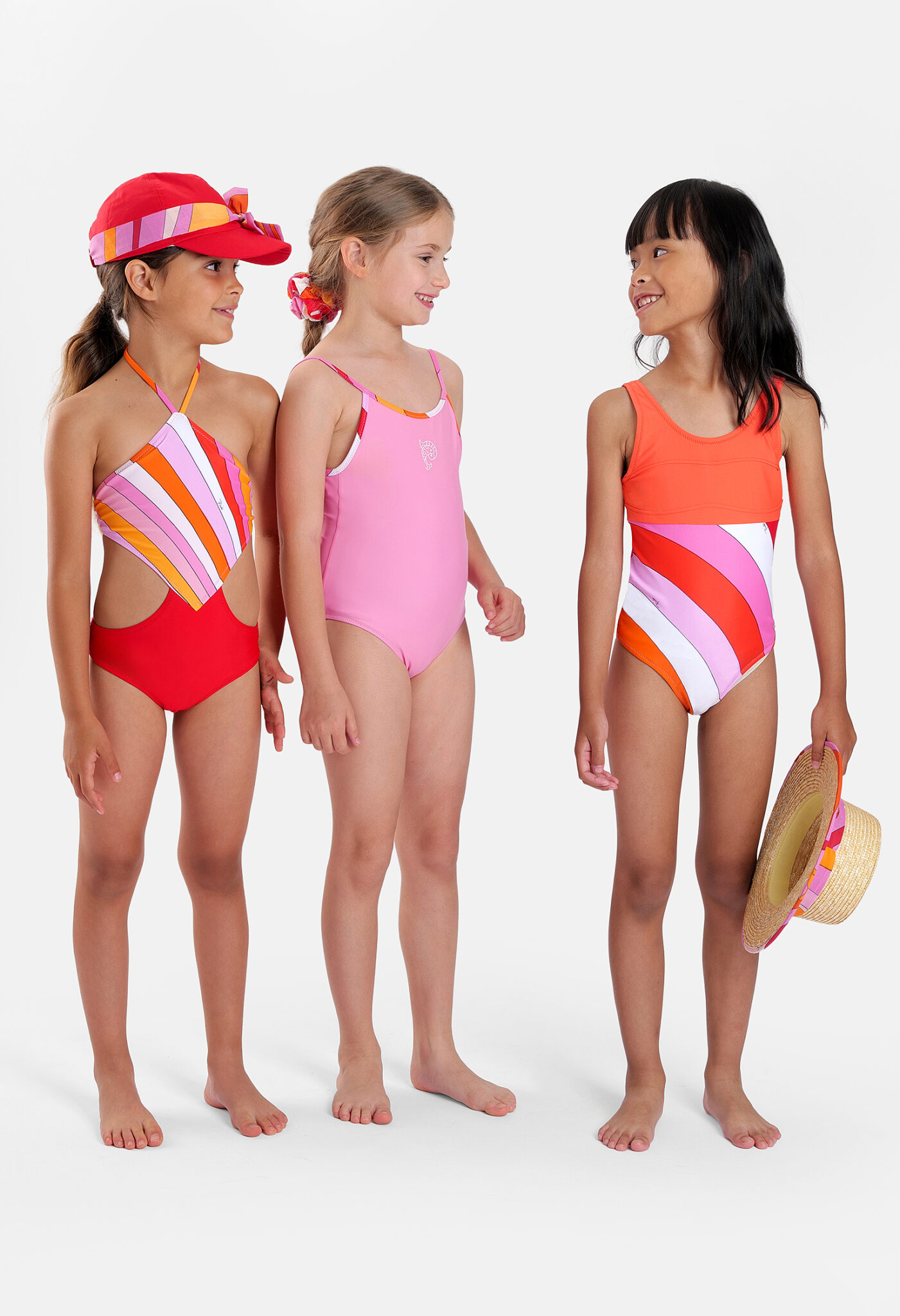 Three girls showcasing Pucci Junior SS26 swimwear collection, featuring colourful one-piece swimsuits and iconic Pucci-inspired graphic prints in pink, orange and lilac tones.