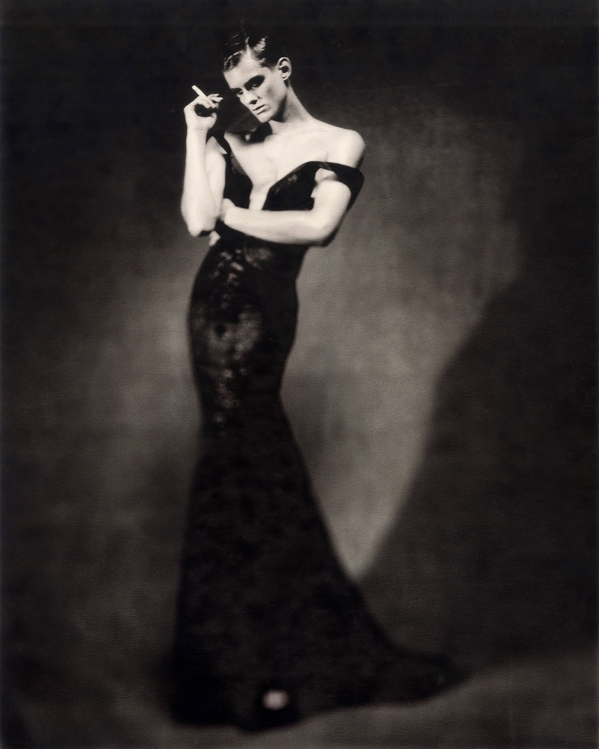 A black-and-white fashion portrait by Paolo Roversi showing a model in a long off-the-shoulder black gown, standing in a graceful pose against a softly lit dark background.
