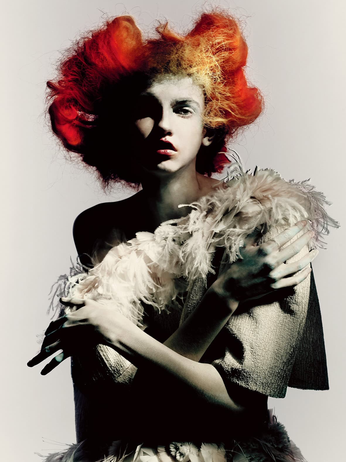 A fashion portrait by Paolo Roversi featuring a model with dramatic red-orange voluminous hair, wrapped in white feathers and sculptural textiles against a minimalist backdrop.