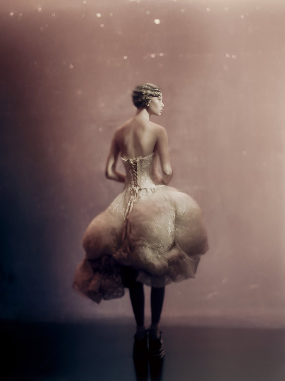 A dreamy fashion portrait by Paolo Roversi depicting a model seen from behind in a sculptural ivory couture dress with a corseted bodice, standing against a soft blush-pink background with a delicate, ethereal glow.