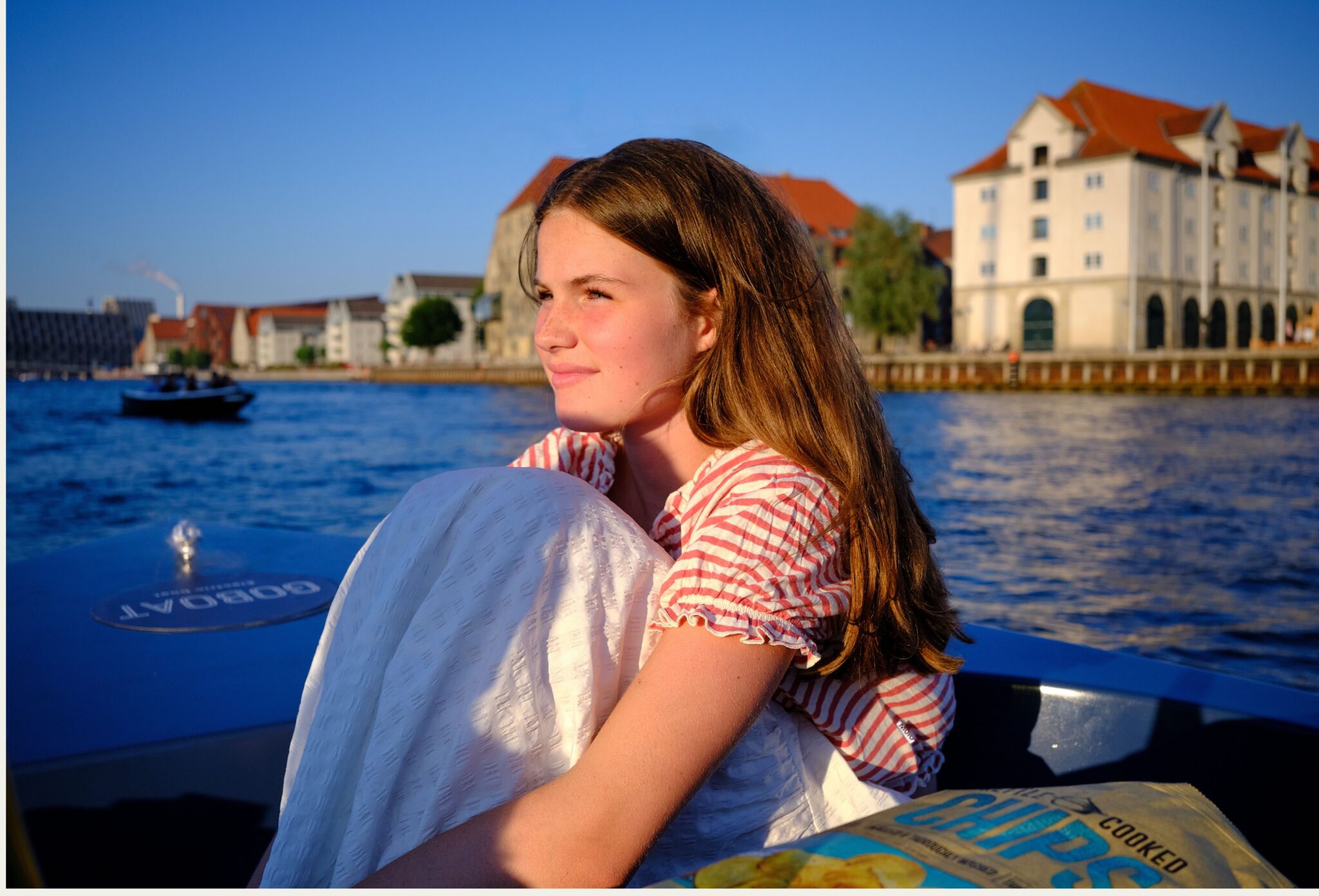 Teen fashion editorial Take Me Back to Tivoli photographed by Erica Allen in Copenhagen, featuring a girl in a striped shirt and white skirt sitting by the water in warm evening light for Scimparello Magazine.