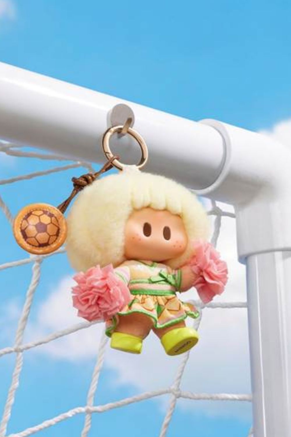 MINISO YOYO Cheer Up Cuteness plush bag charm hanging from a football goal, featuring fluffy blonde hair, pink pom-poms and a sporty outfit designed as a collectible backpack accessory.