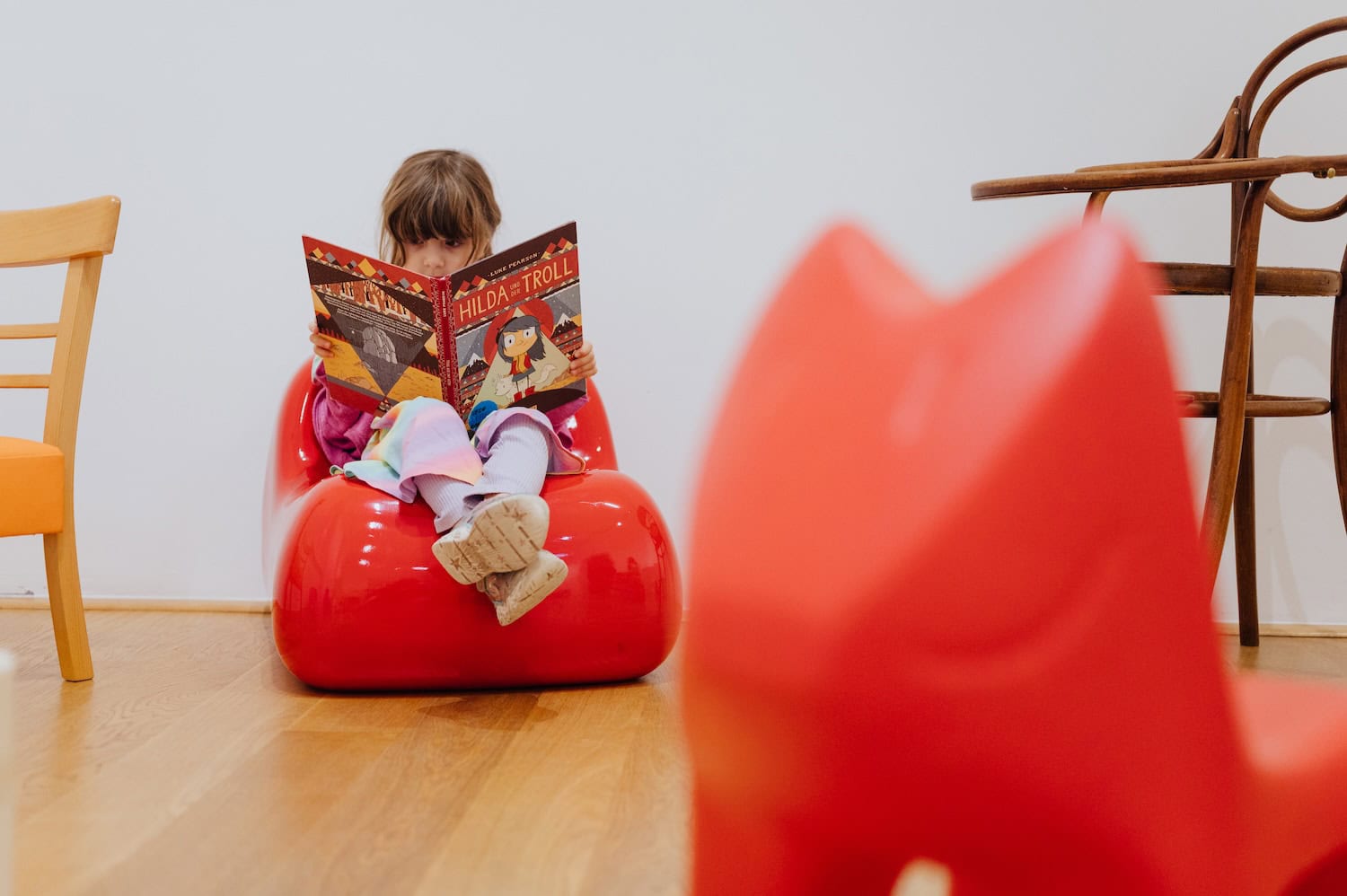Young child reading a book while sitting on a bright red designer children's chair at the Mini Furniture: Chairs for Children exhibition at MK&G Hamburg.