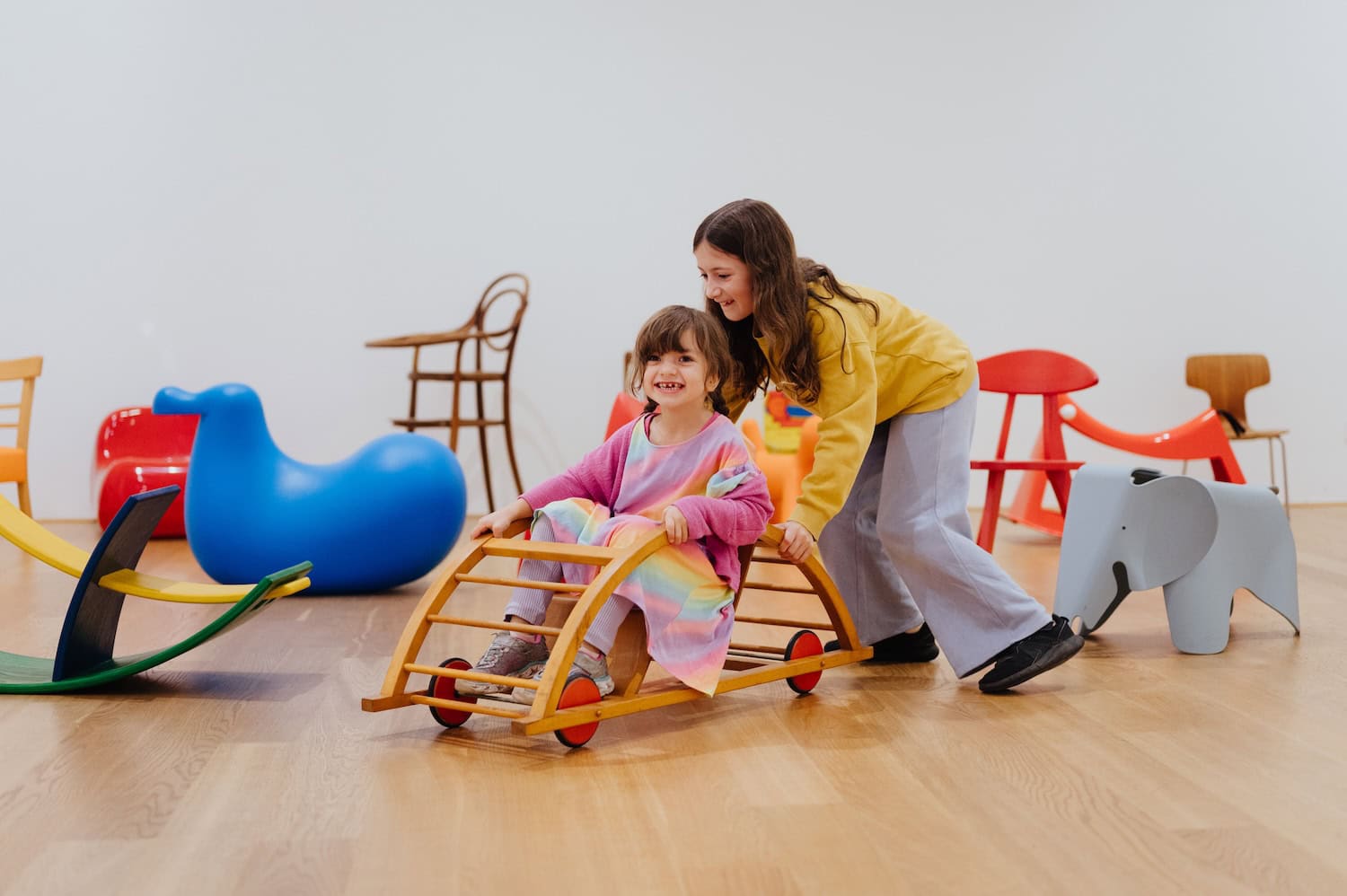 Two children play with a rolling wooden chair surrounded by iconic children's furniture during the interactive Mini Furniture: Chairs for Children exhibition at MK&G Hamburg.