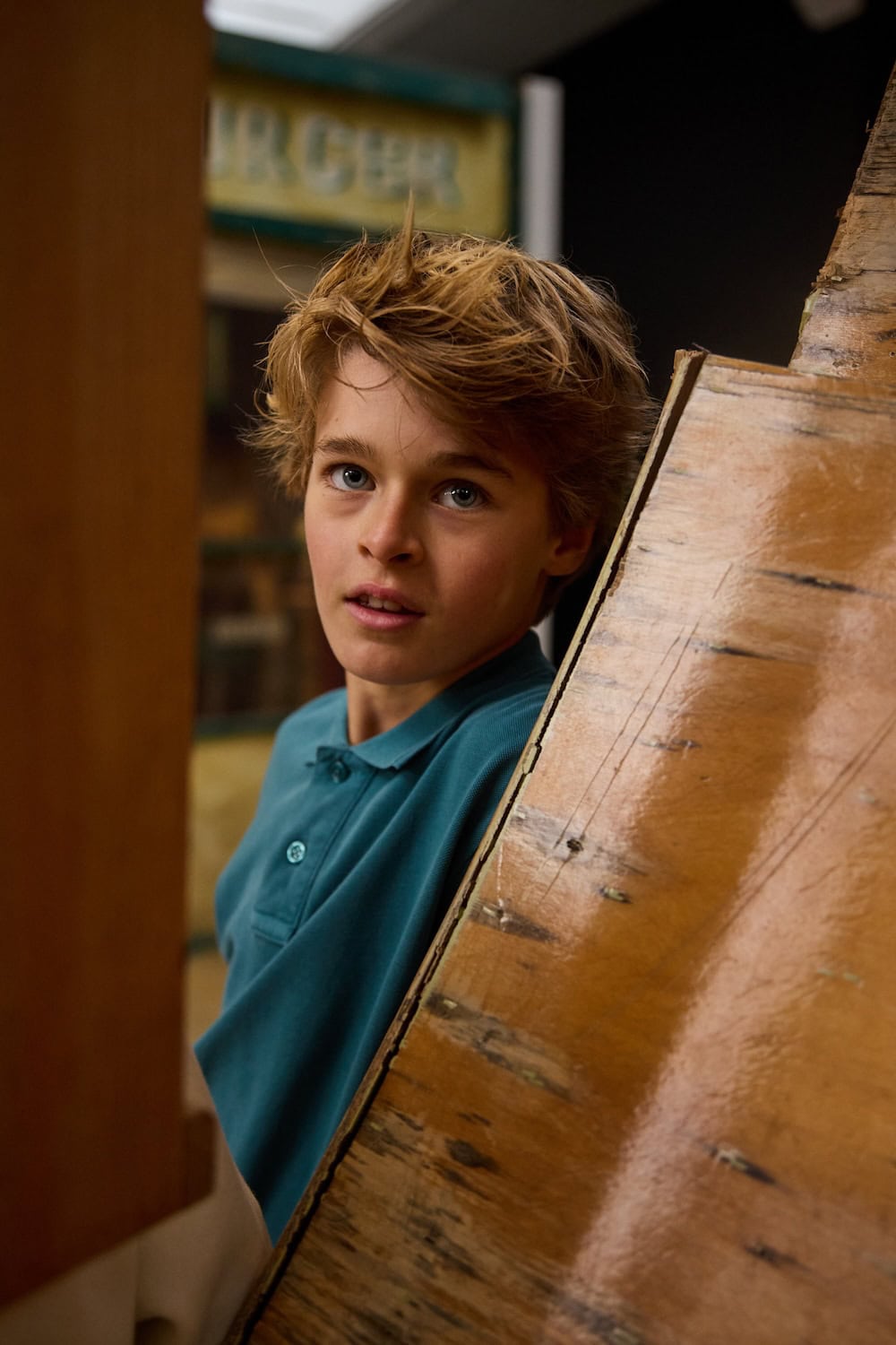Boy in teal polo shirt peeking from behind a wooden structure inside a vintage industrial building, wearing casual autumn childrenswear.
