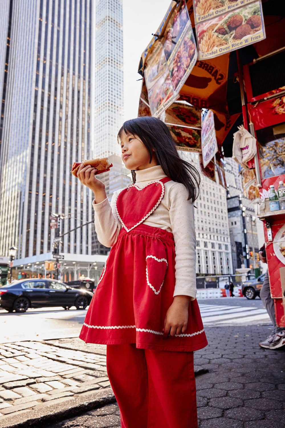 Girl eating a hot dog in Manhattan wearing a red heart dress from the Bebe Organic AW26 collection.