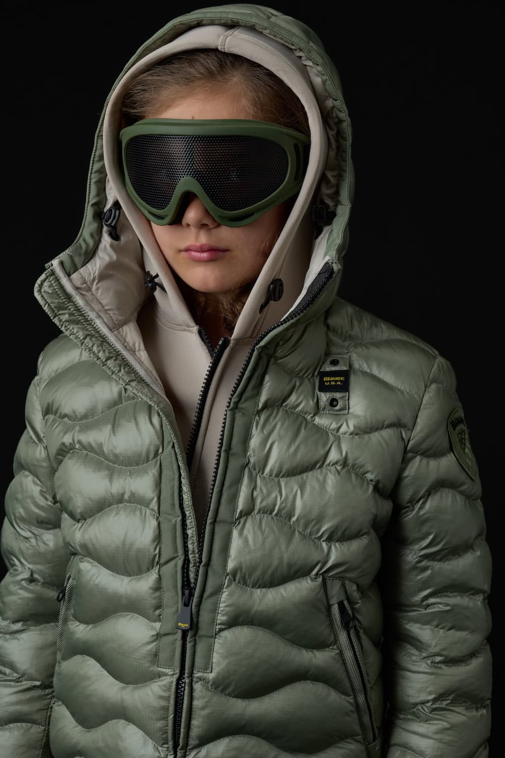 Close-up of a girl wearing a sage green Blauer Junior hooded quilted puffer jacket layered with a beige sweatshirt and oversized ski goggles.