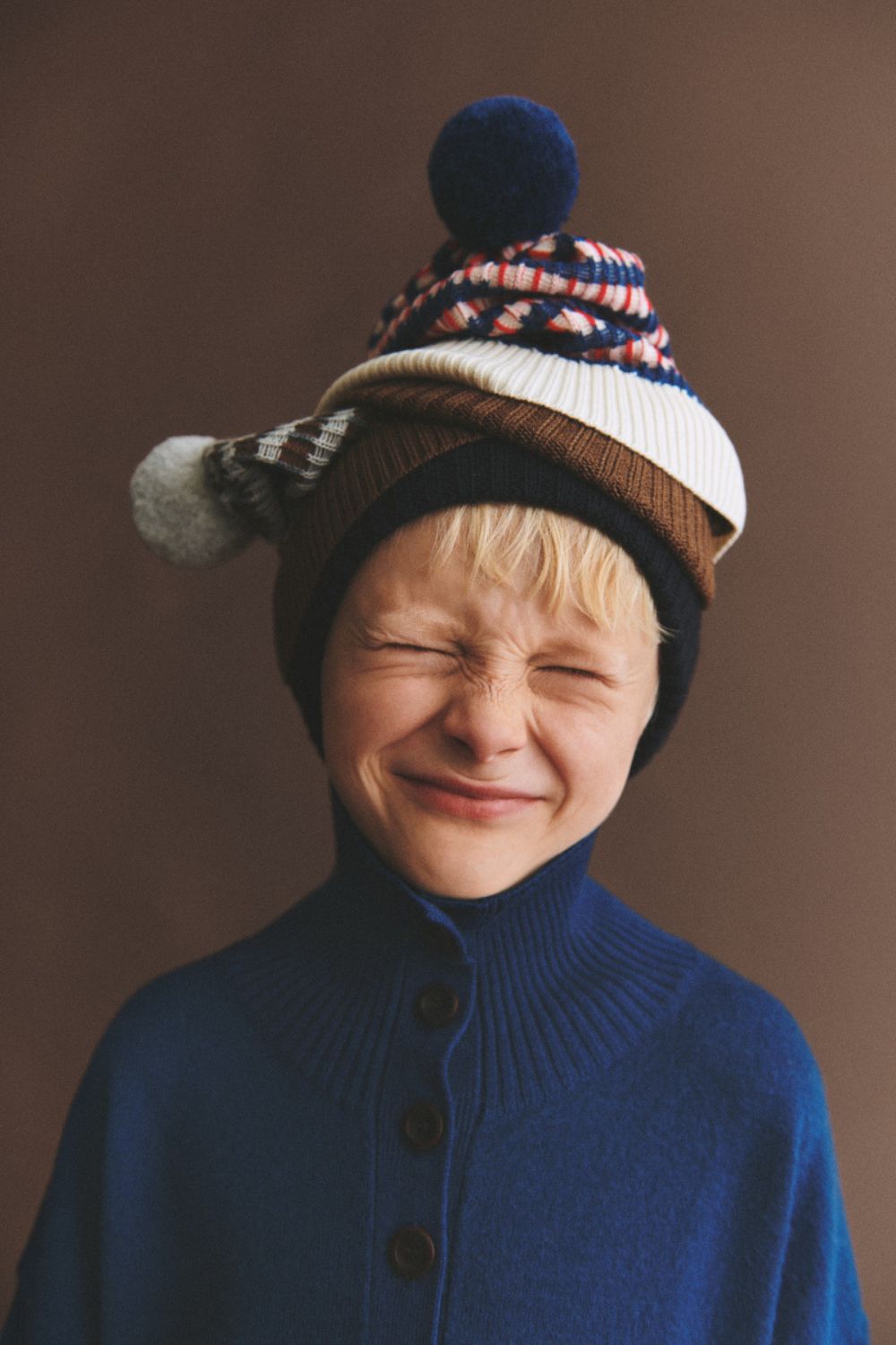 Boy wearing a royal blue high-neck knitted sweater and stacked colorful knit hats from the FUB Kids AW26 collection.